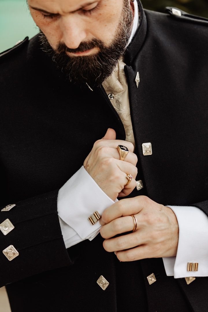 Bearded groom in black Scottish military jacket adjusting gold cufflinks on white dress shirt.