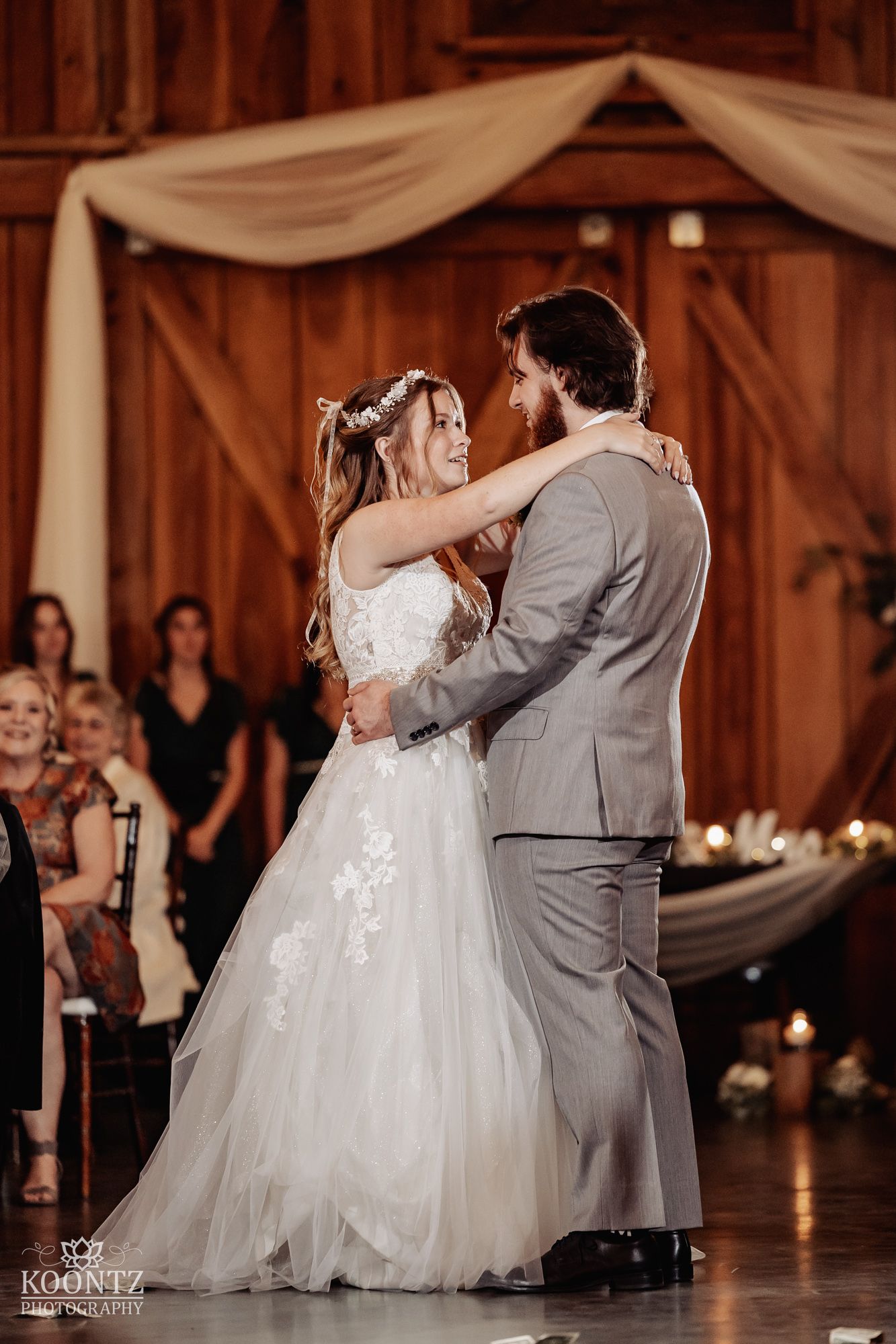 Bride and groom share their first dance in a rustic barn wedding venue with draped fabric backdrop.