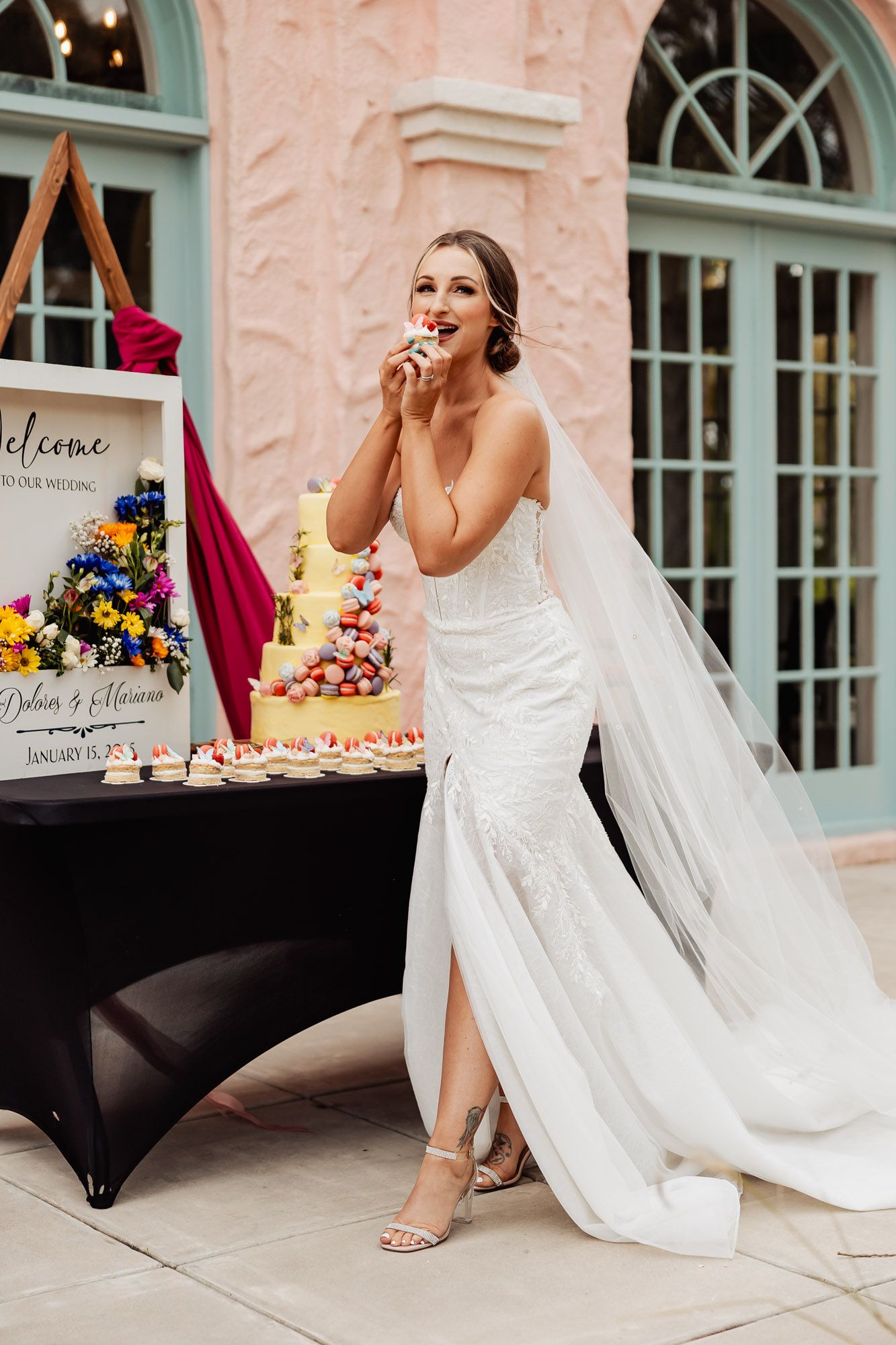 Bride in fitted white gown with veil enjoys a treat at a colorful dessert table outside a pink venue.