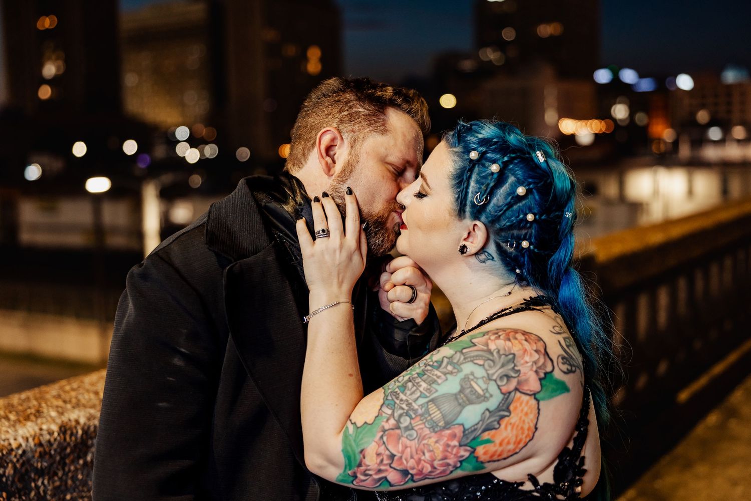 Tattooed couple sharing romantic kiss at night with city lights bokeh background at wedding.