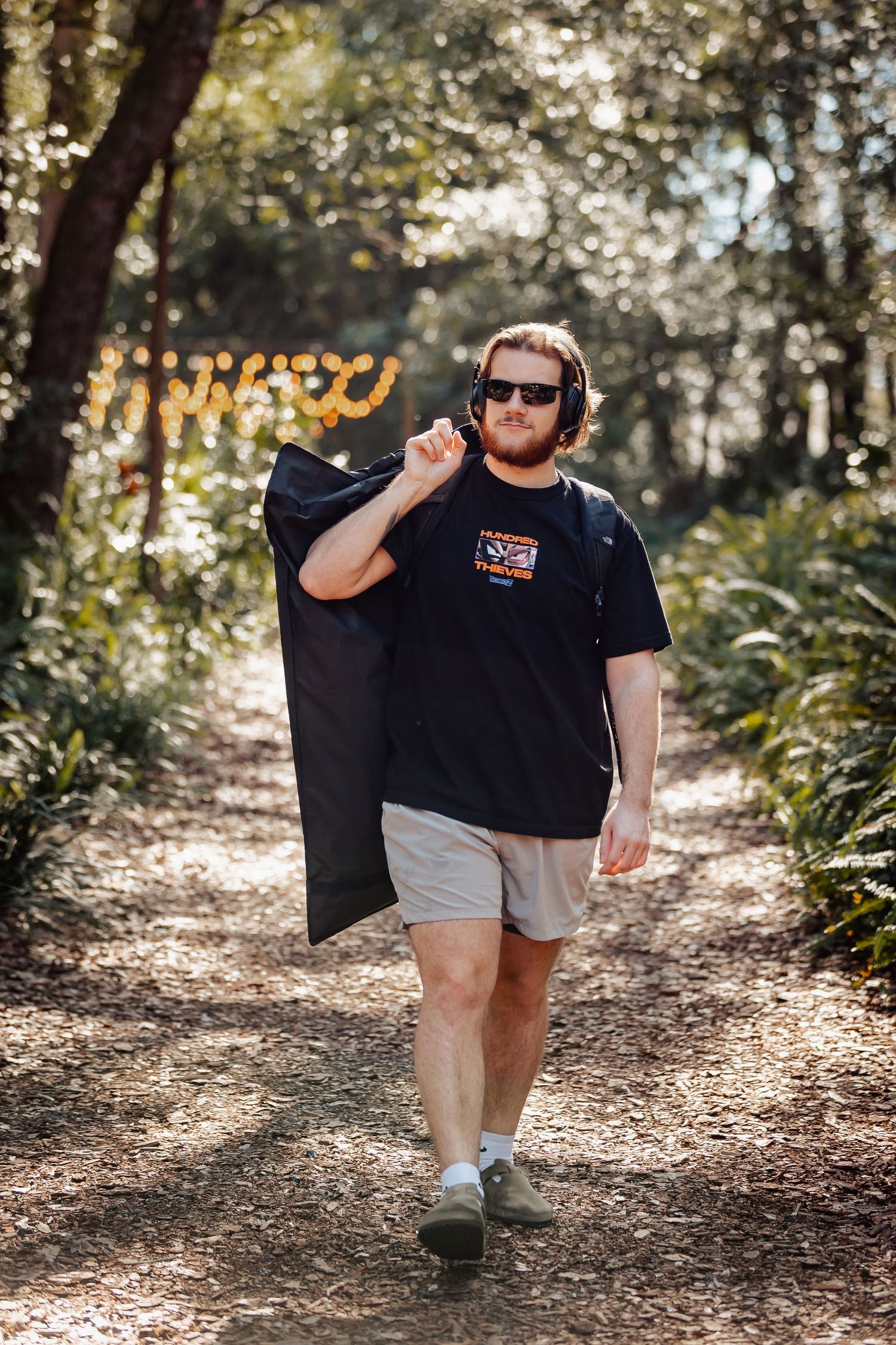 Person in casual attire carrying a garment bag while walking along a sunlit woodland path.