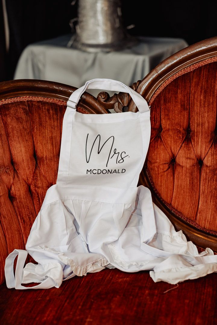 White apron with 'Mrs McDonald' text displayed on vintage red tufted chair.