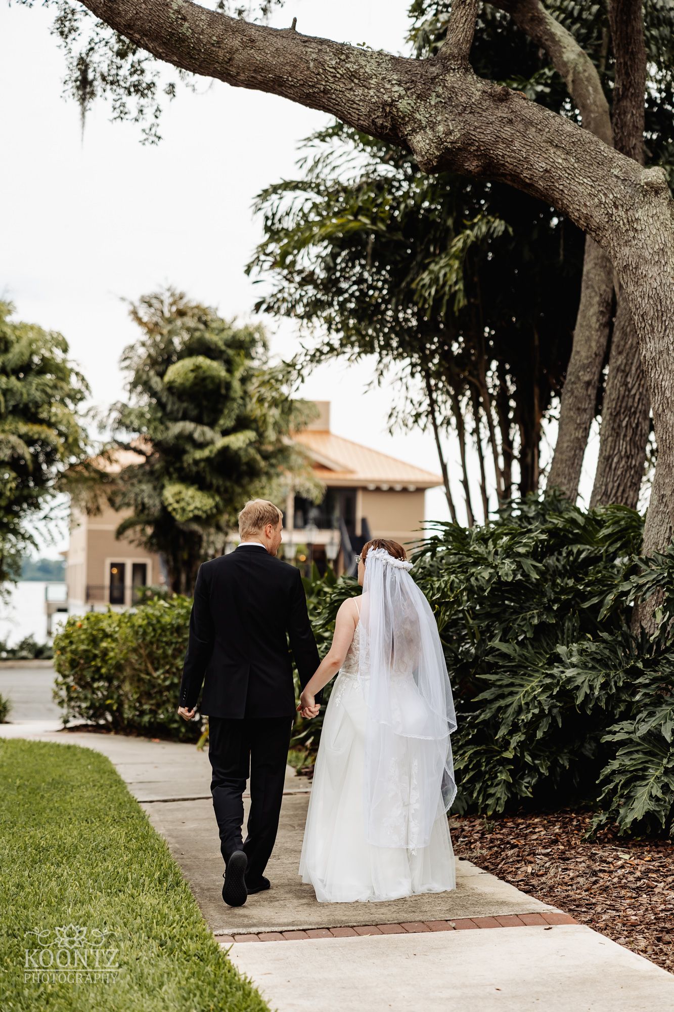 Couple walking along a garden path with lush tropical landscaping and Spanish-style architecture in the background.