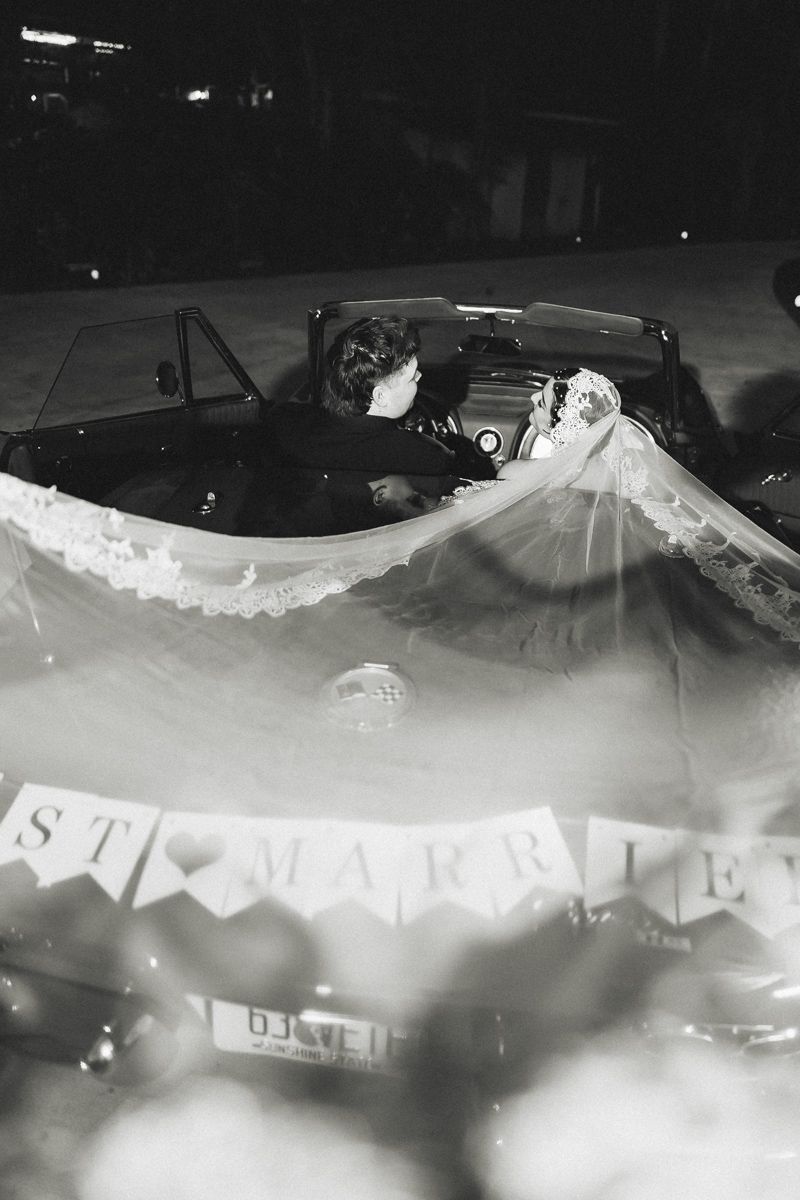 Black and white photo of newlyweds kissing in a vintage convertible with Just Married banner.