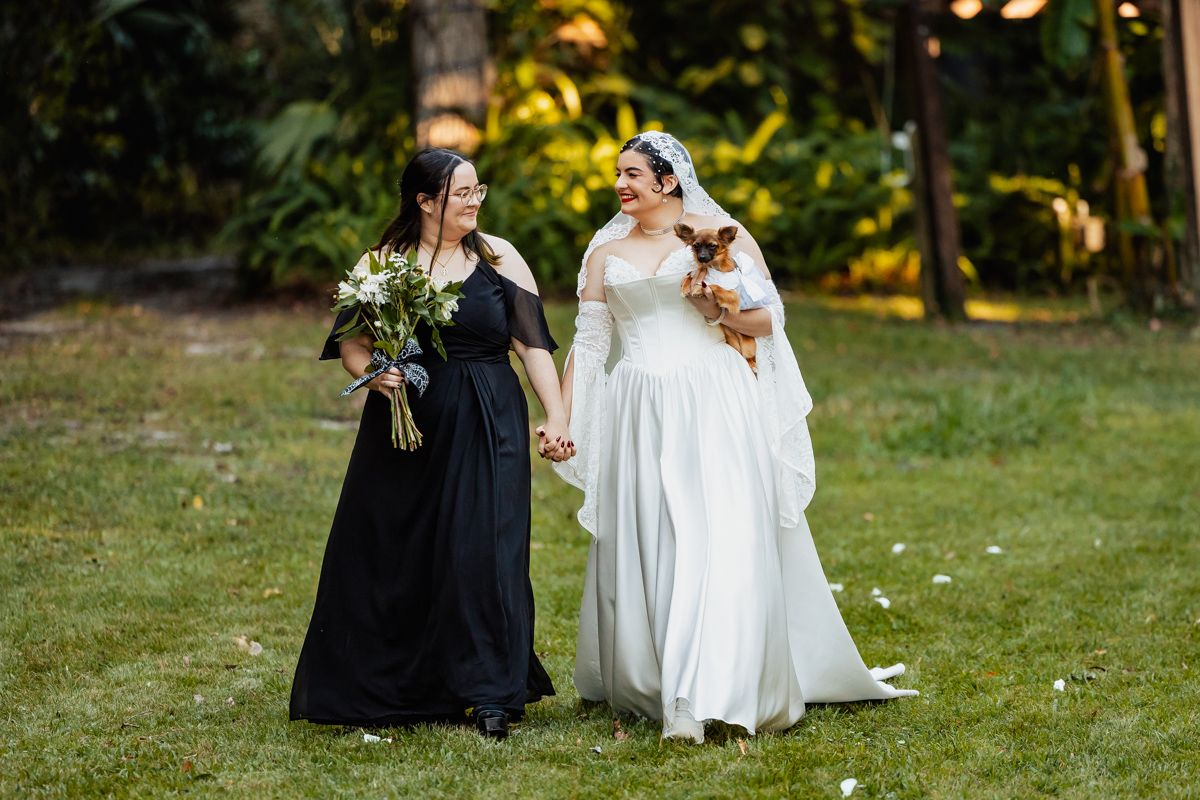 Bride in white gown and bridesmaid in black dress walking together holding bouquet and small dog.