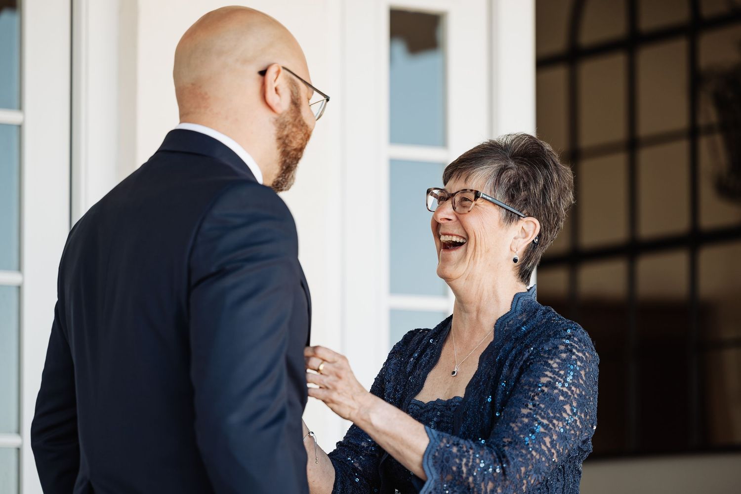 Older woman in blue lace dress laughing joyfully with bald groom in navy suit at wedding.