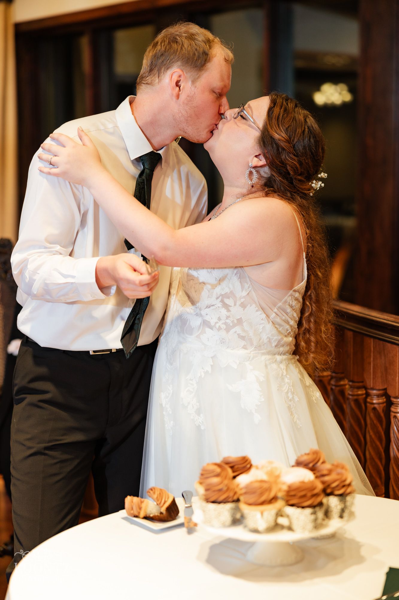 Newlyweds share a romantic kiss next to a tiered display of desserts at their wedding reception.