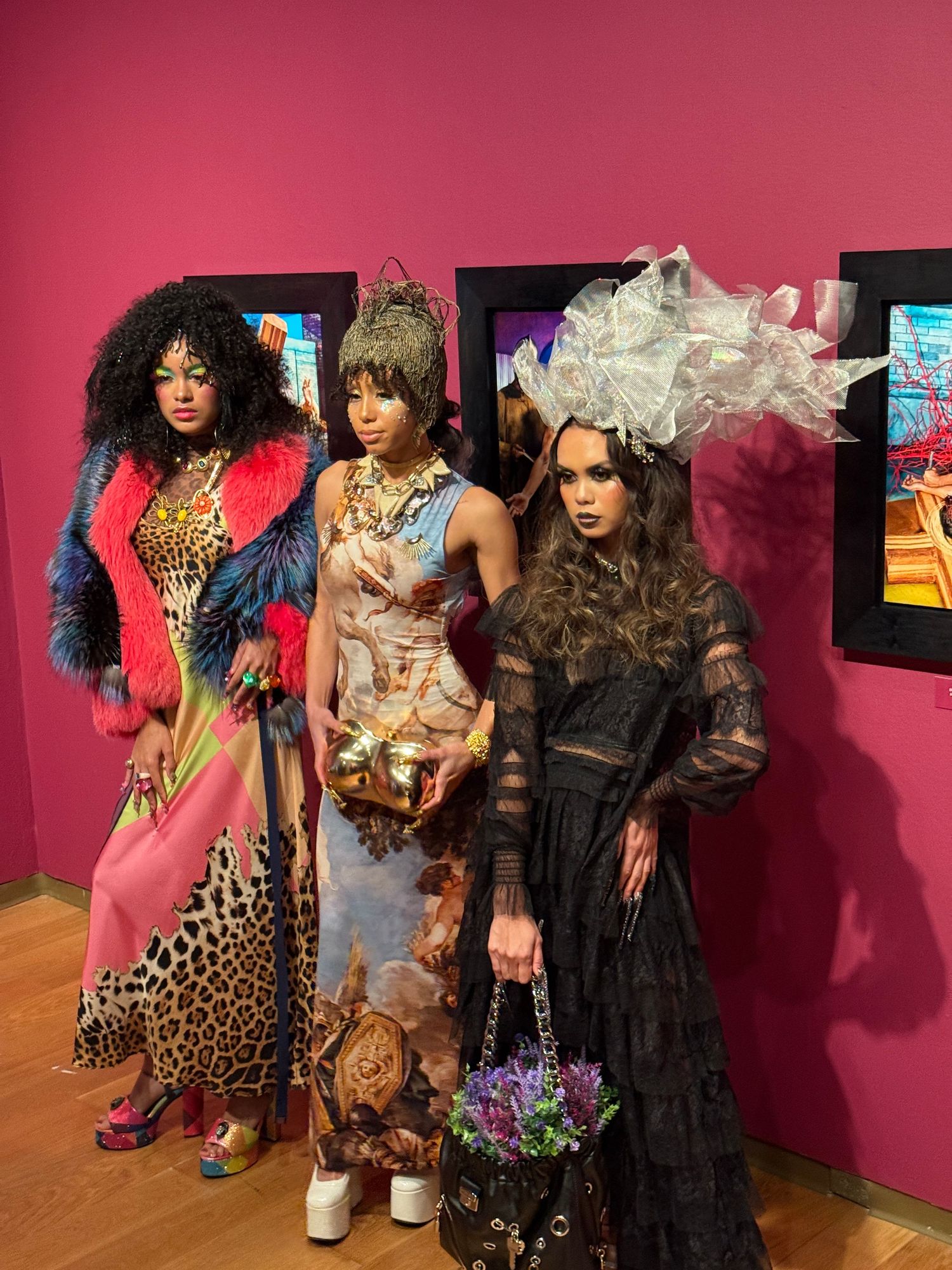 Fashion models showcase eclectic outfits with animal prints, lace, and bold colors against a pink backdrop in an art gallery.