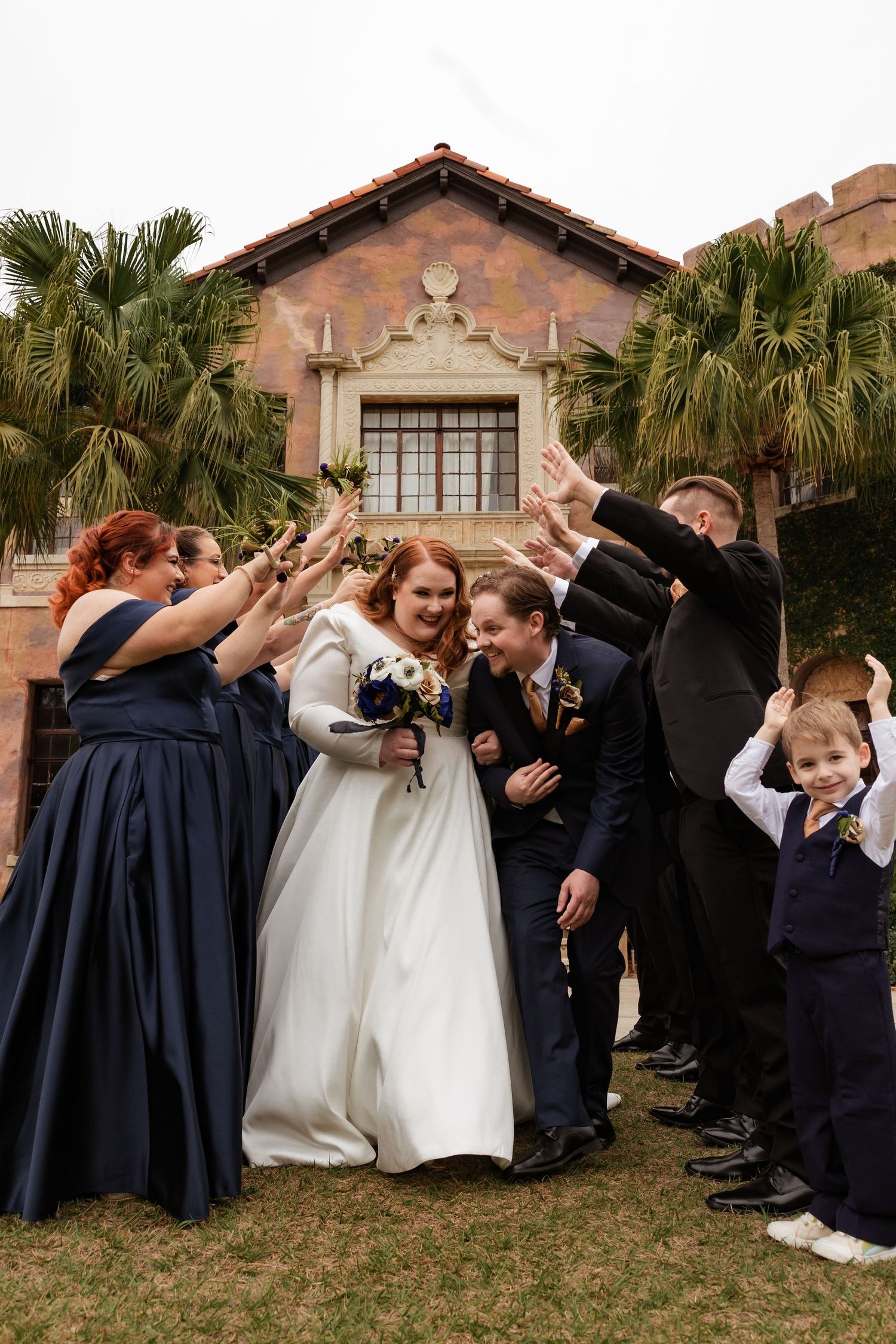 Bride and groom walk through celebratory arch formed by cheering wedding party outside historic venue.
