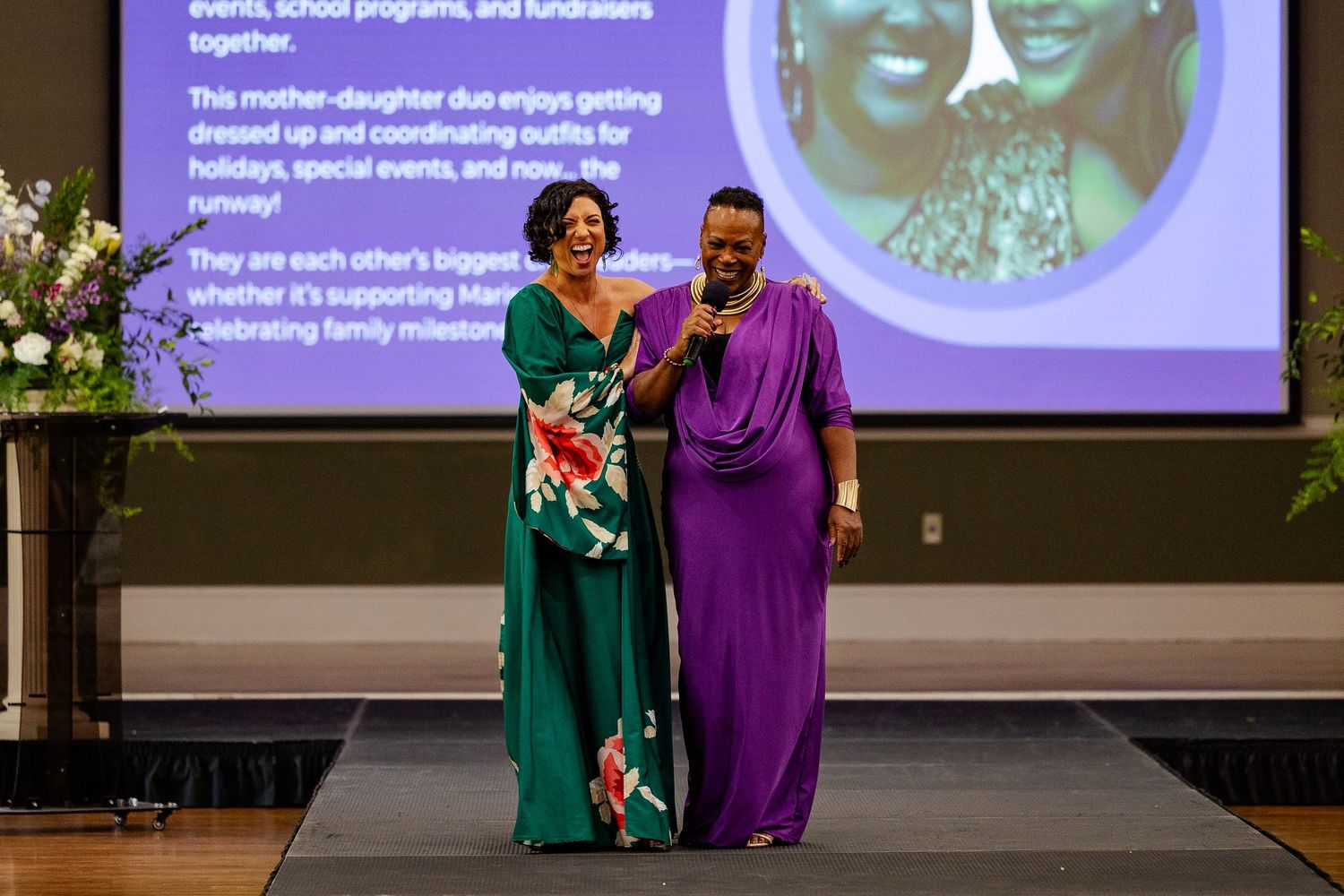 Two women in elegant gowns pose together on stage at a formal fashion event.