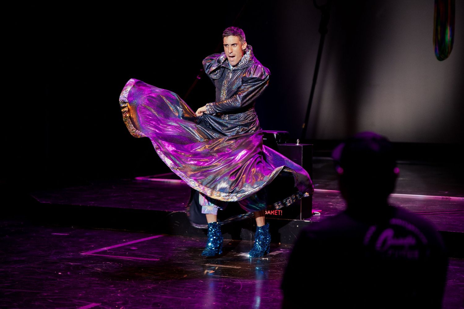 Stage performer twirling in large iridescent purple cape under colorful spotlight on dark stage.