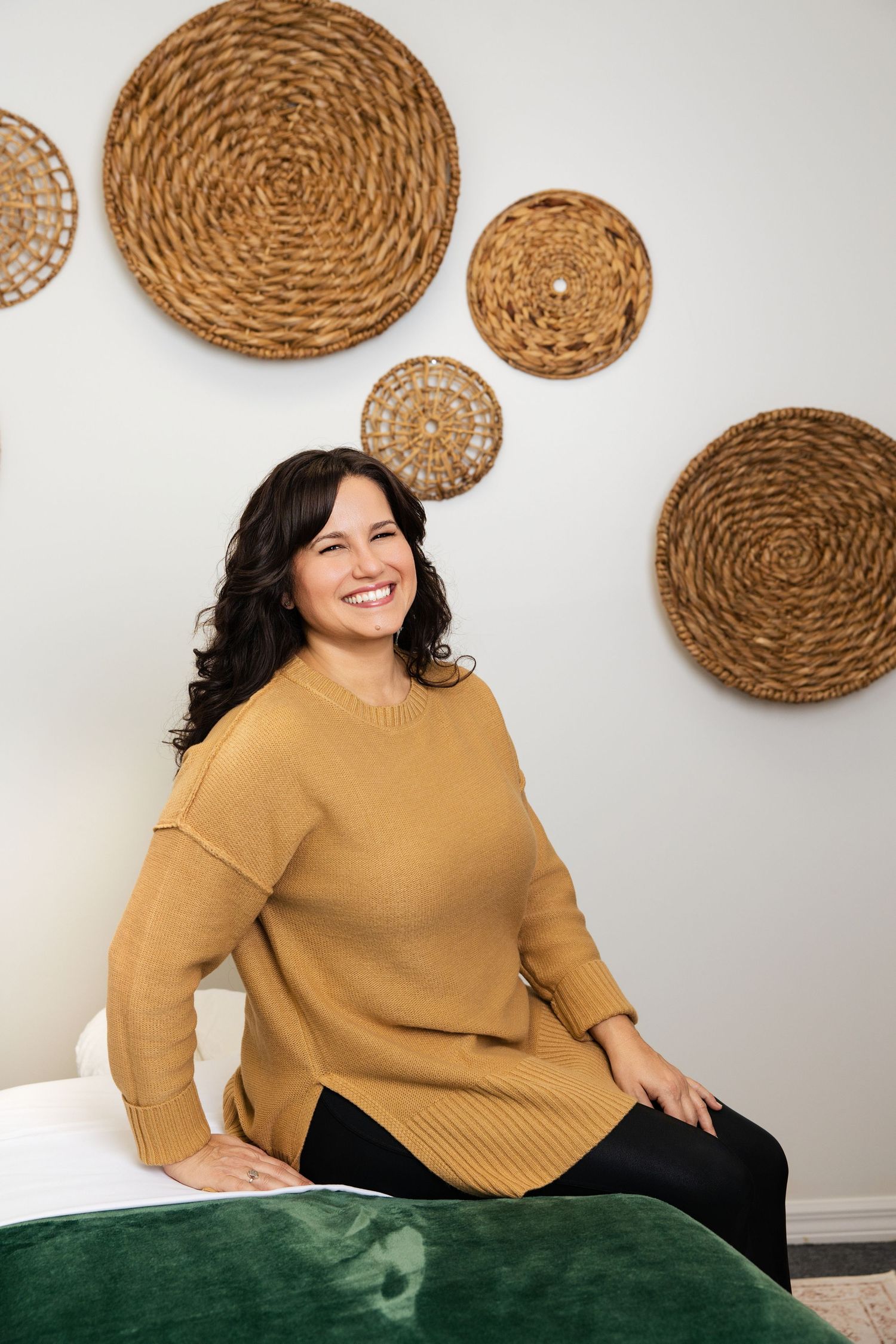 Smiling woman in mustard yellow sweater sitting on a massage table with wicker wall decor behind her.