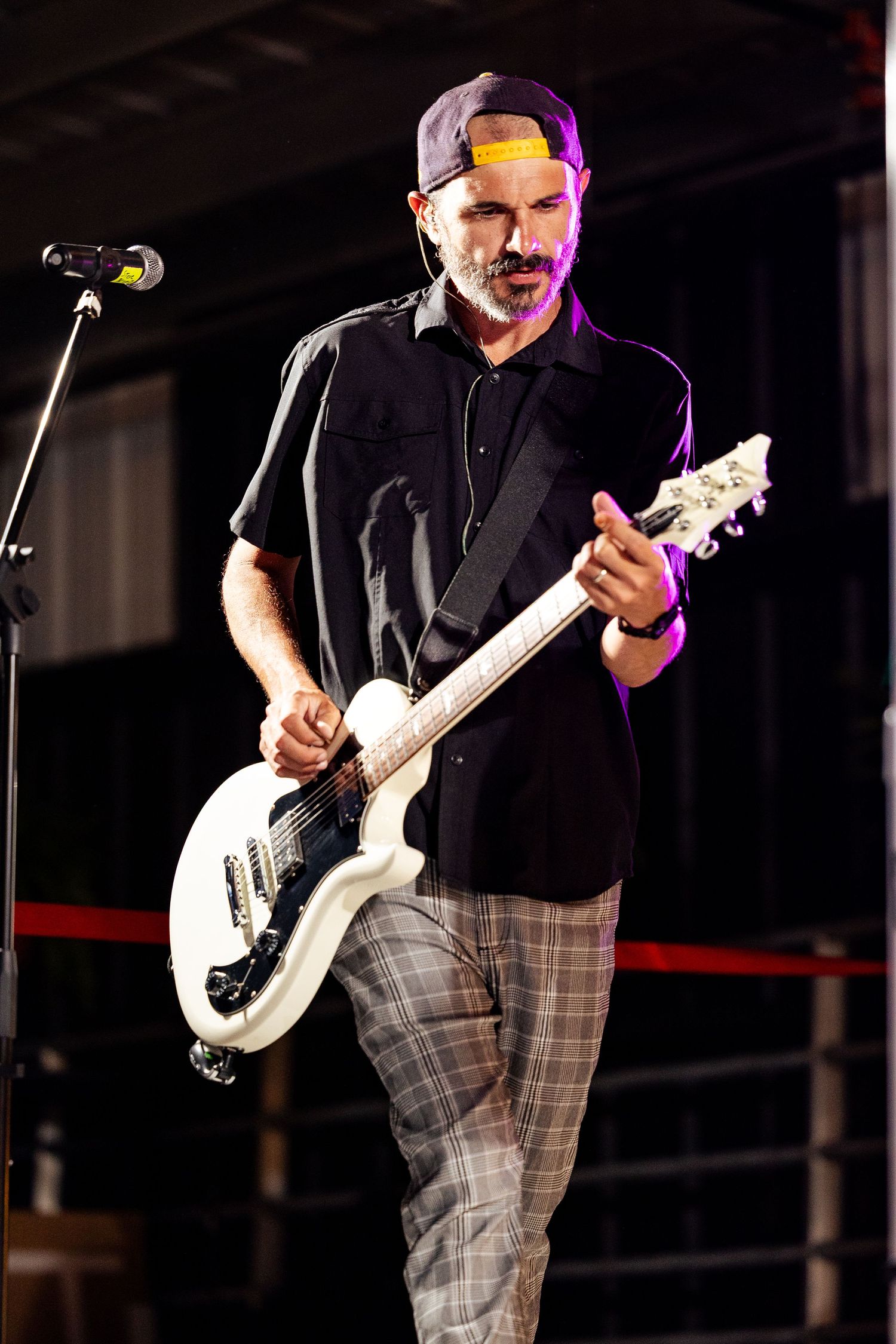 A bearded male guitarist in a black shirt and cap plays a white electric guitar on a dimly lit stage.