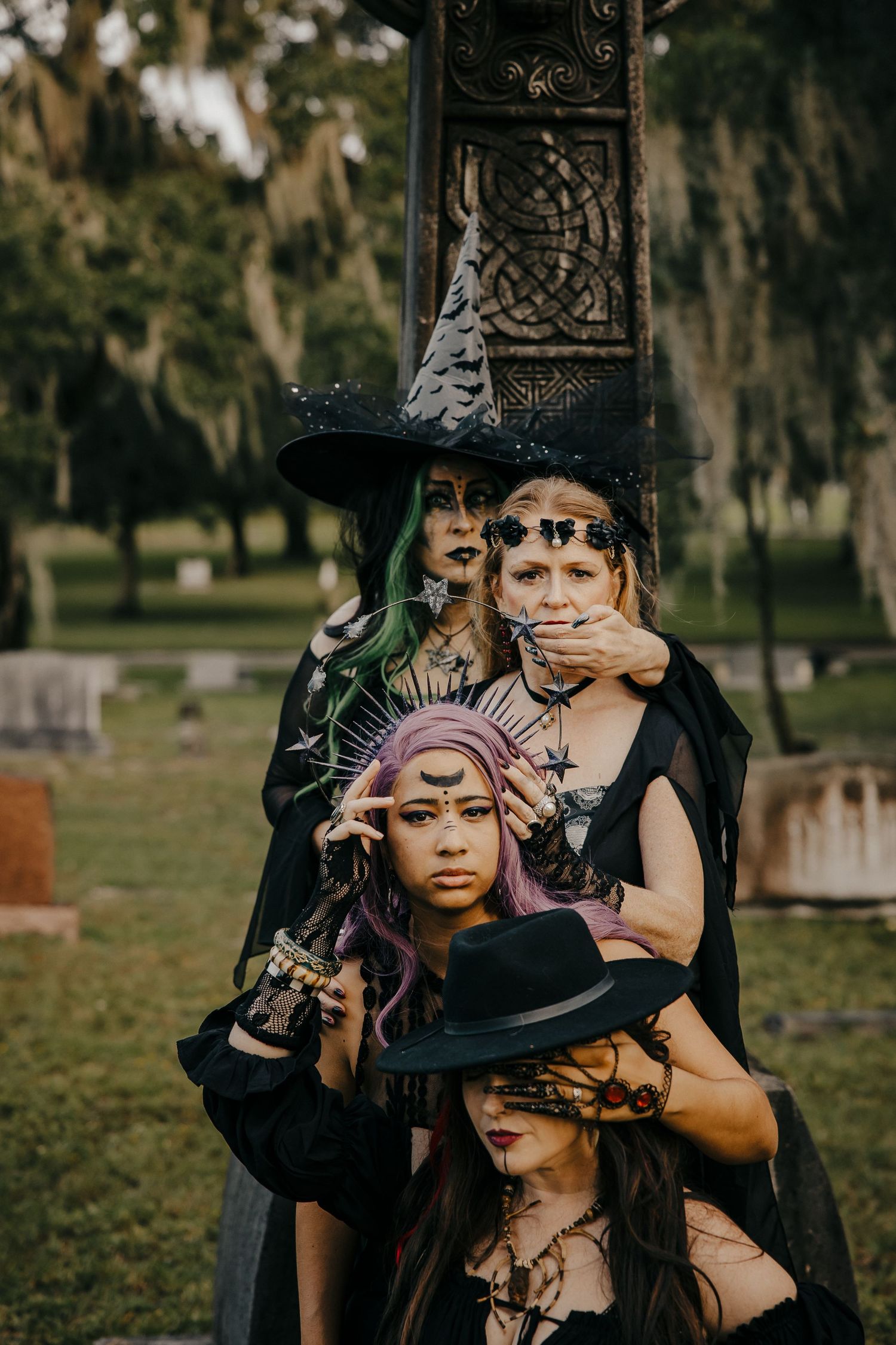 Four women in witch costumes with dramatic face paint pose together beside a Celtic cross gravestone.
