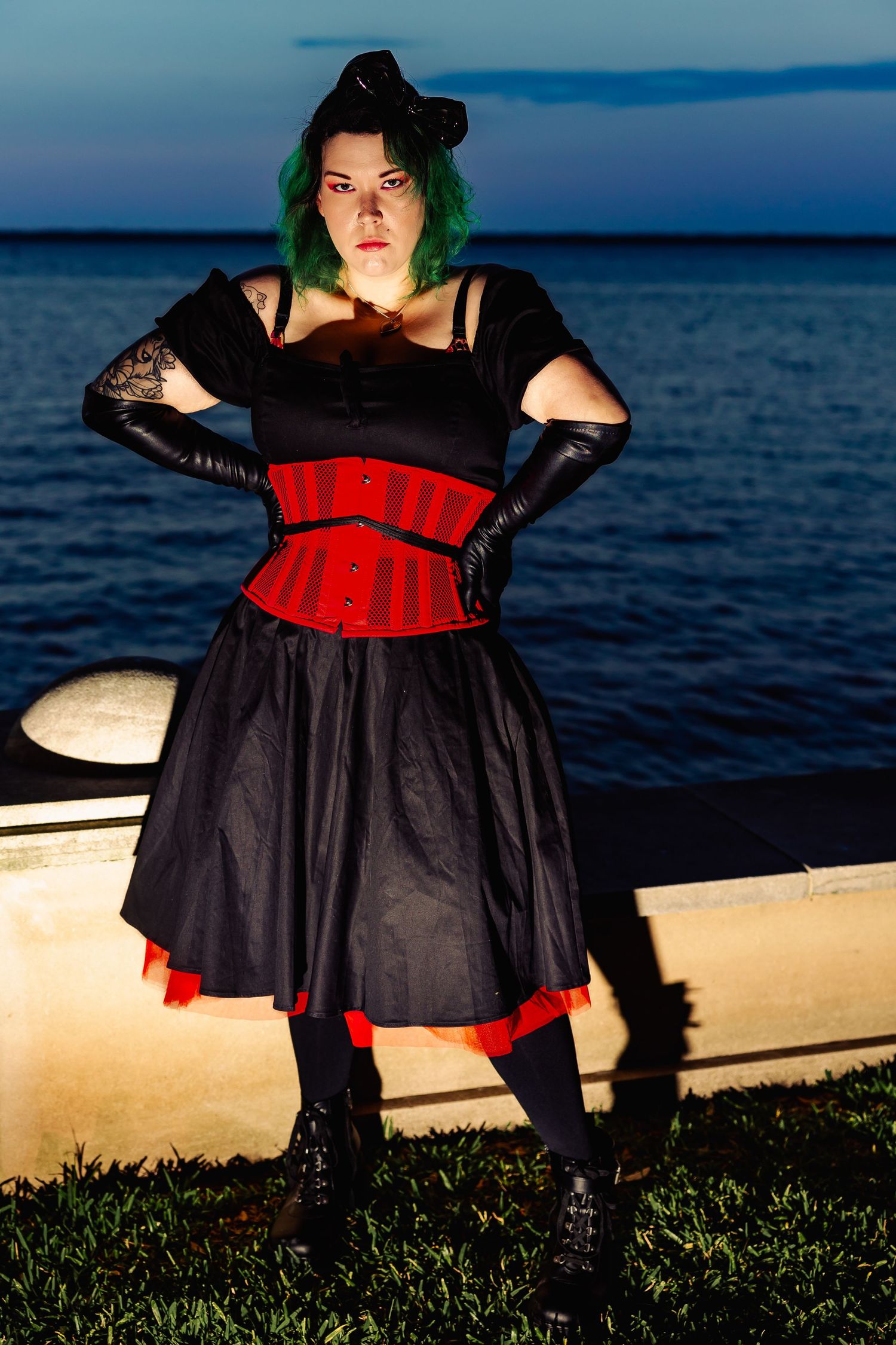 Woman in gothic black and red corset dress with green hair stands dramatically near waterfront at night.