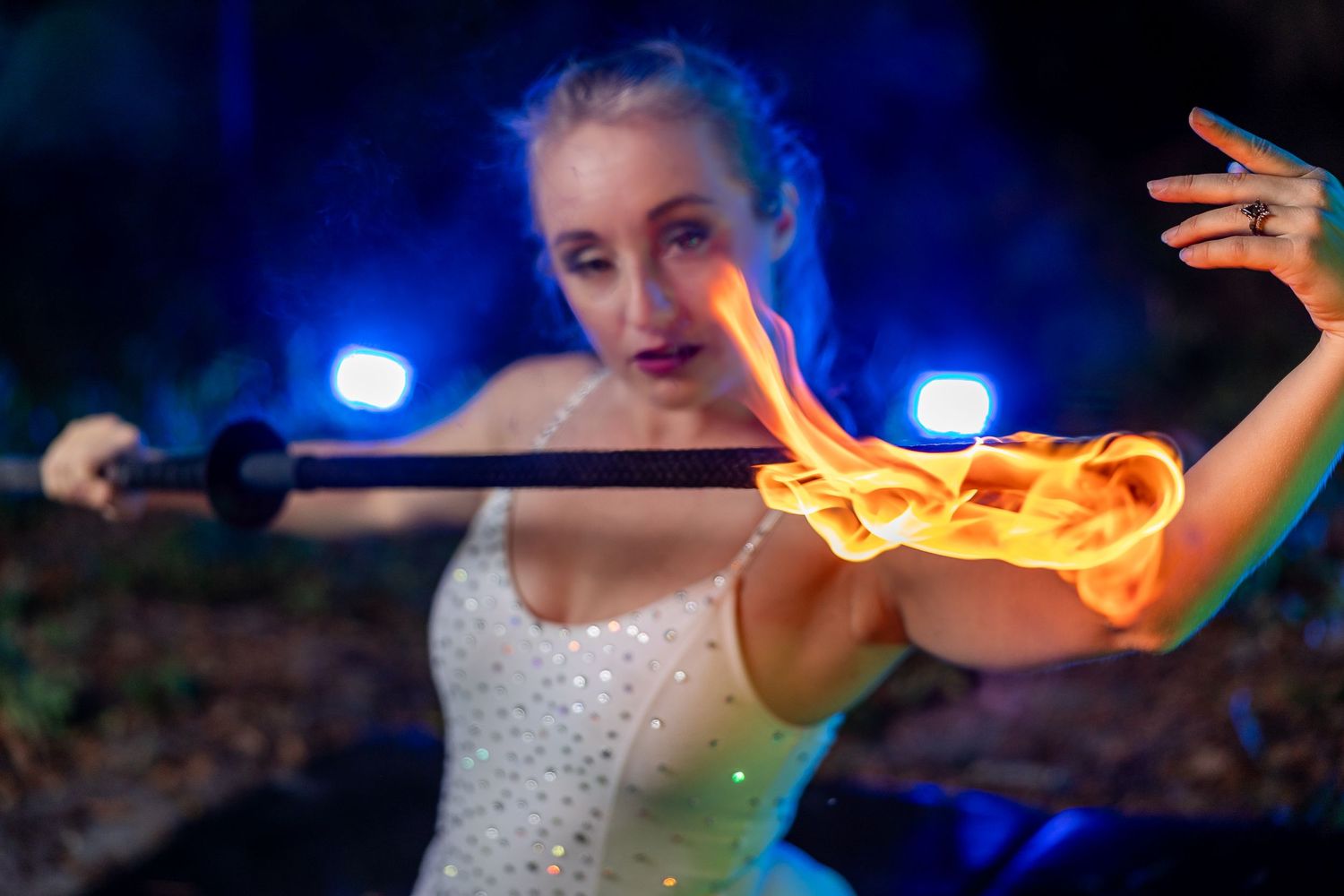 Blonde female fire dancer extending a flaming staff with blue stage lighting in outdoor night setting.