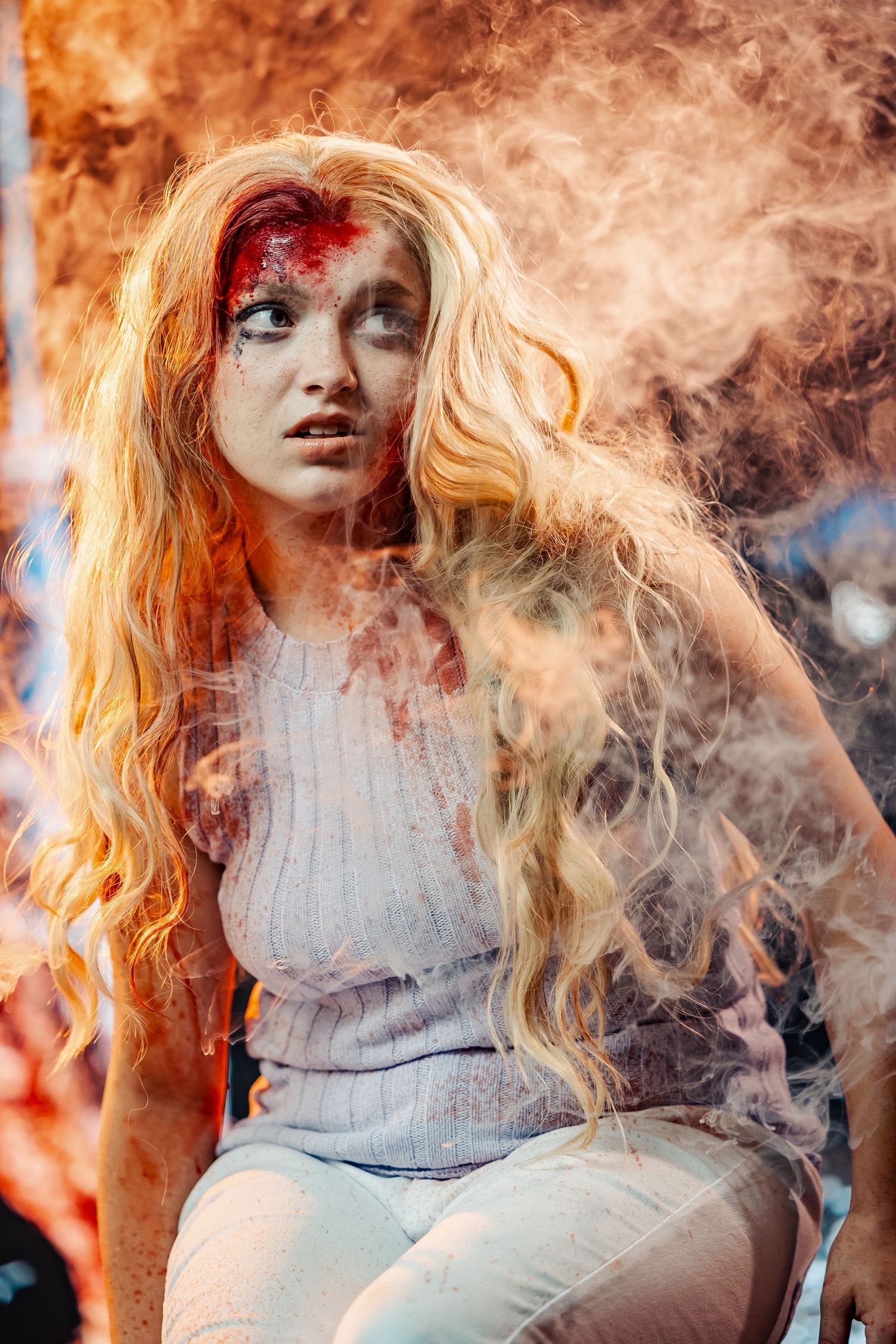 Young blonde woman with horror makeup and fake blood wounds surrounded by smoke and fire effects.