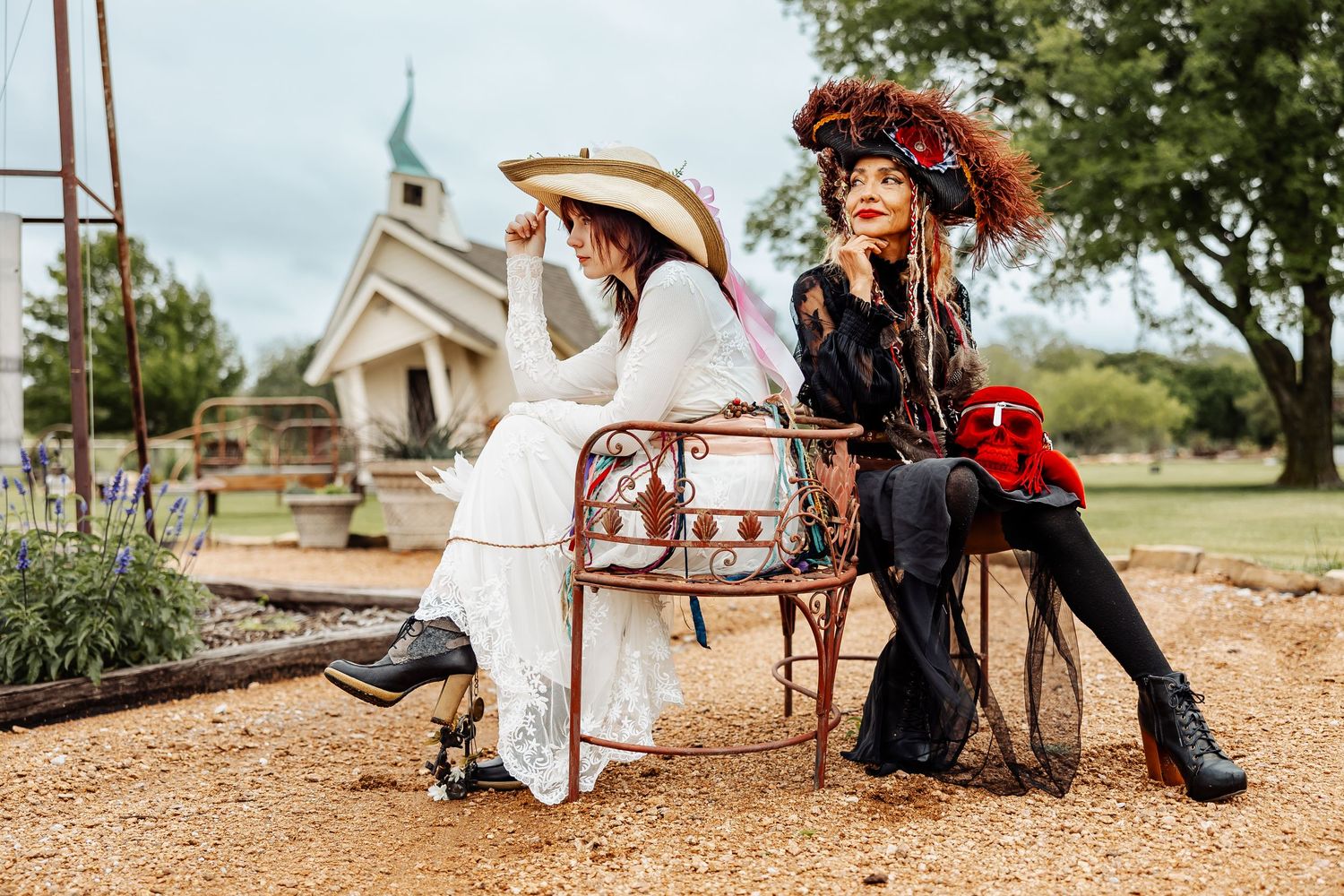 Two women in vintage costumes sit on iron chairs near a white chapel in a garden setting.