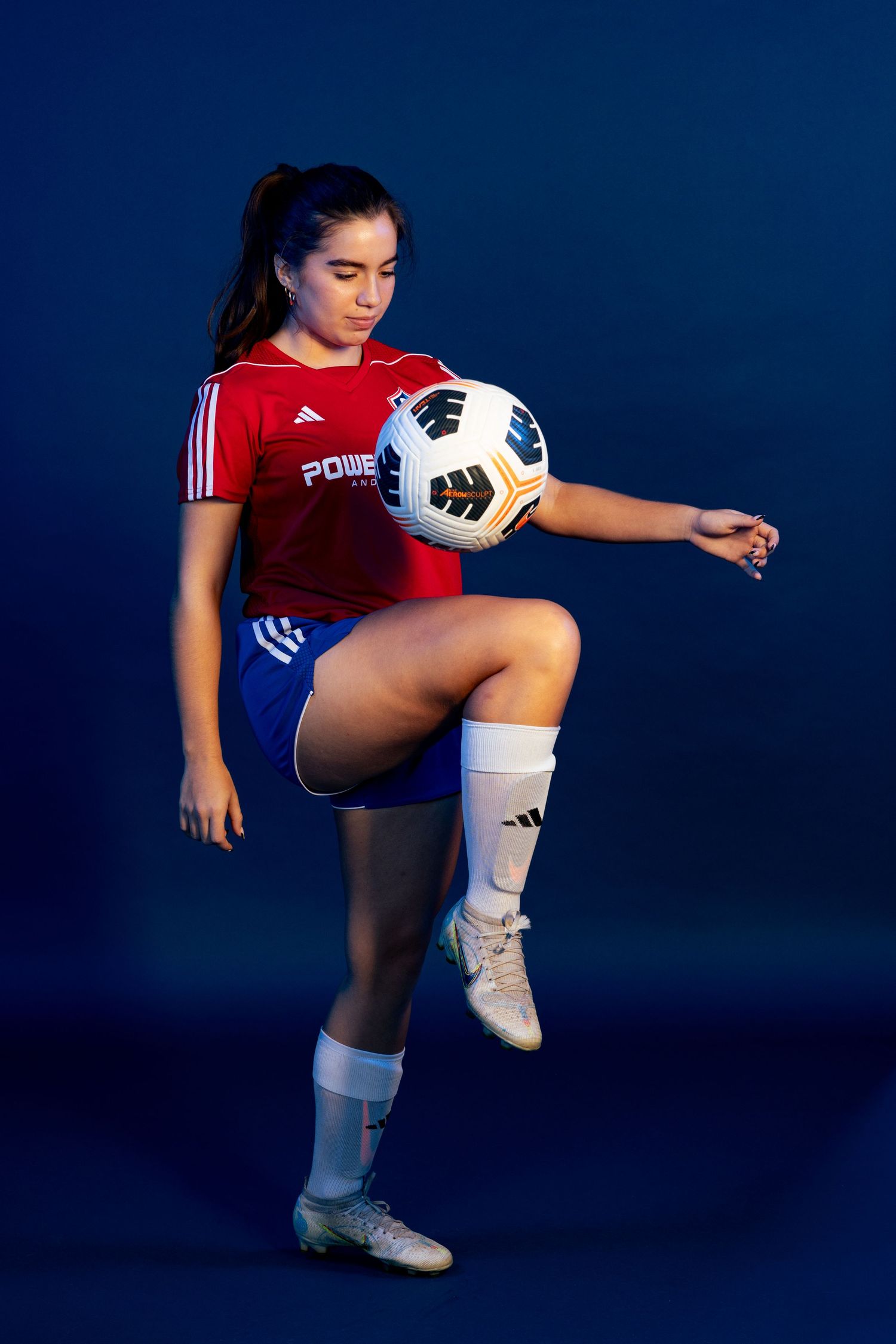 Young female soccer player in red jersey and blue shorts balancing a soccer ball on her knee against a dark blue background.