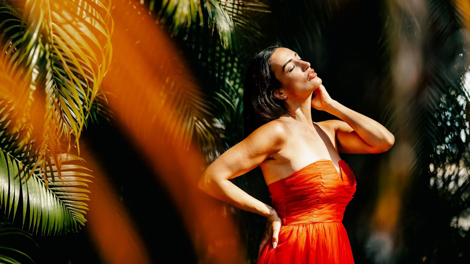 Woman in red strapless dress poses gracefully among lush tropical palm leaves in warm golden sunlight.