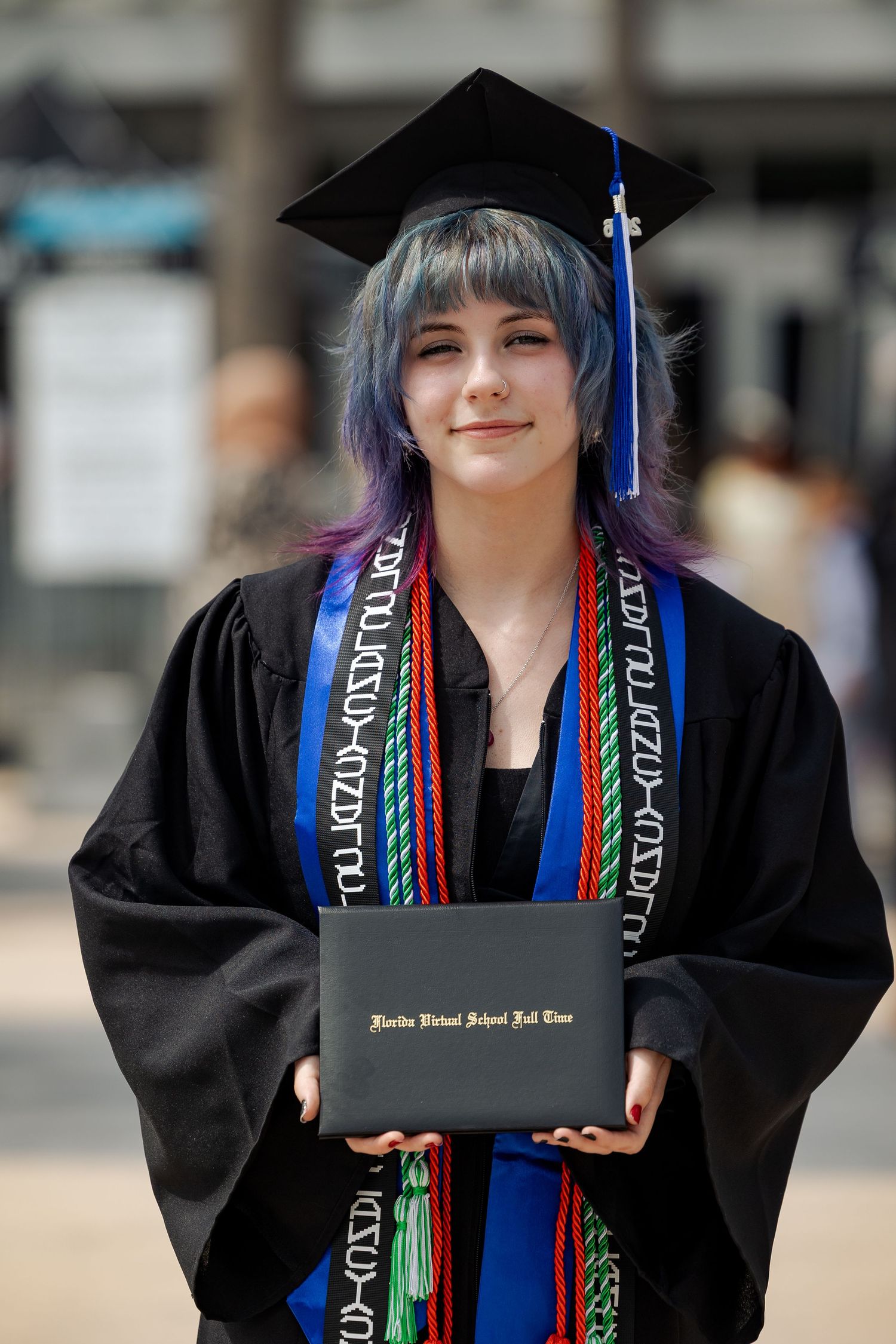 Smiling graduate with blue-purple hair holds diploma in black cap, gown, and colorful honor cords.