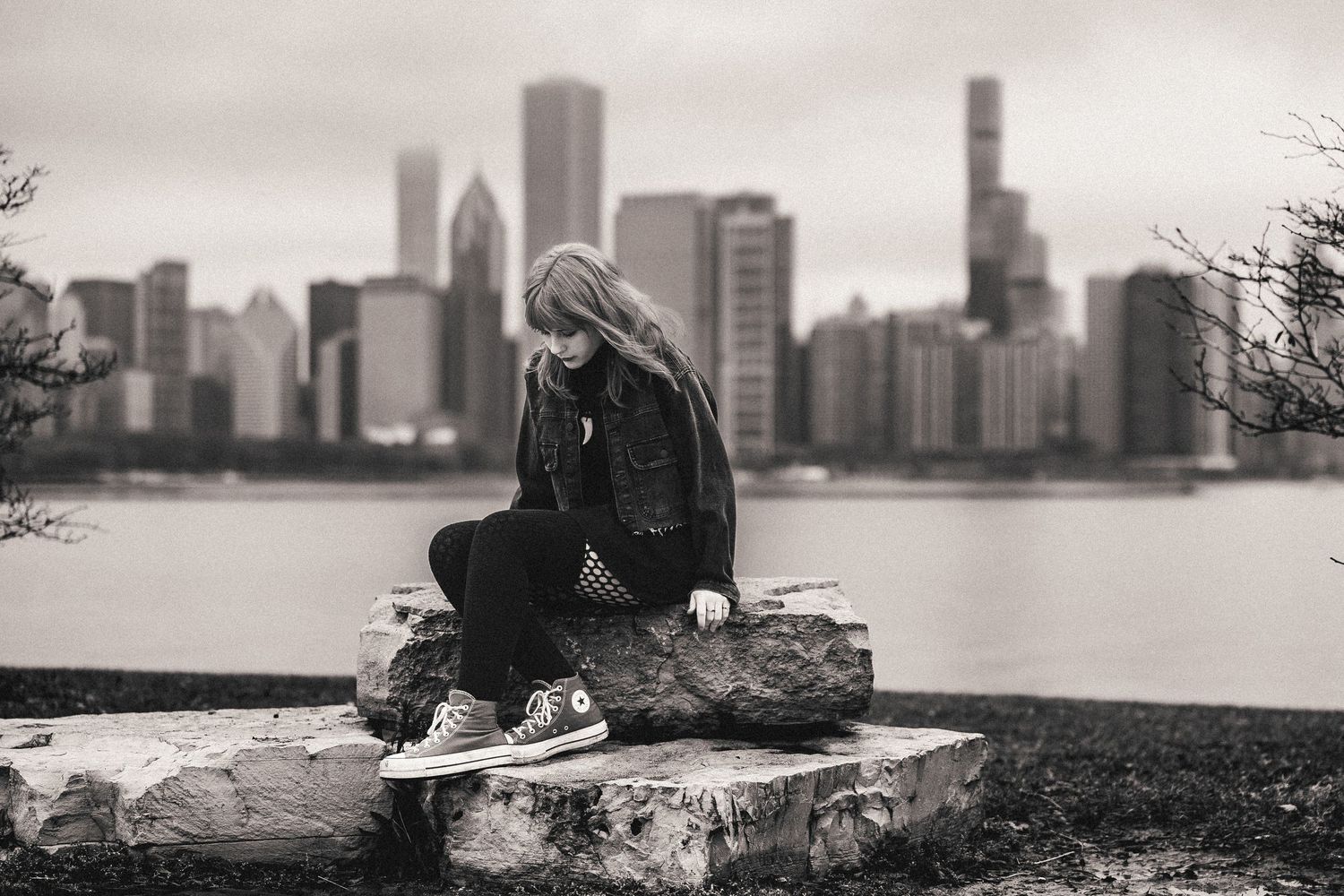 Black and white photo of a young woman sitting on rocks with a skateboard, city skyline and river in the background.
