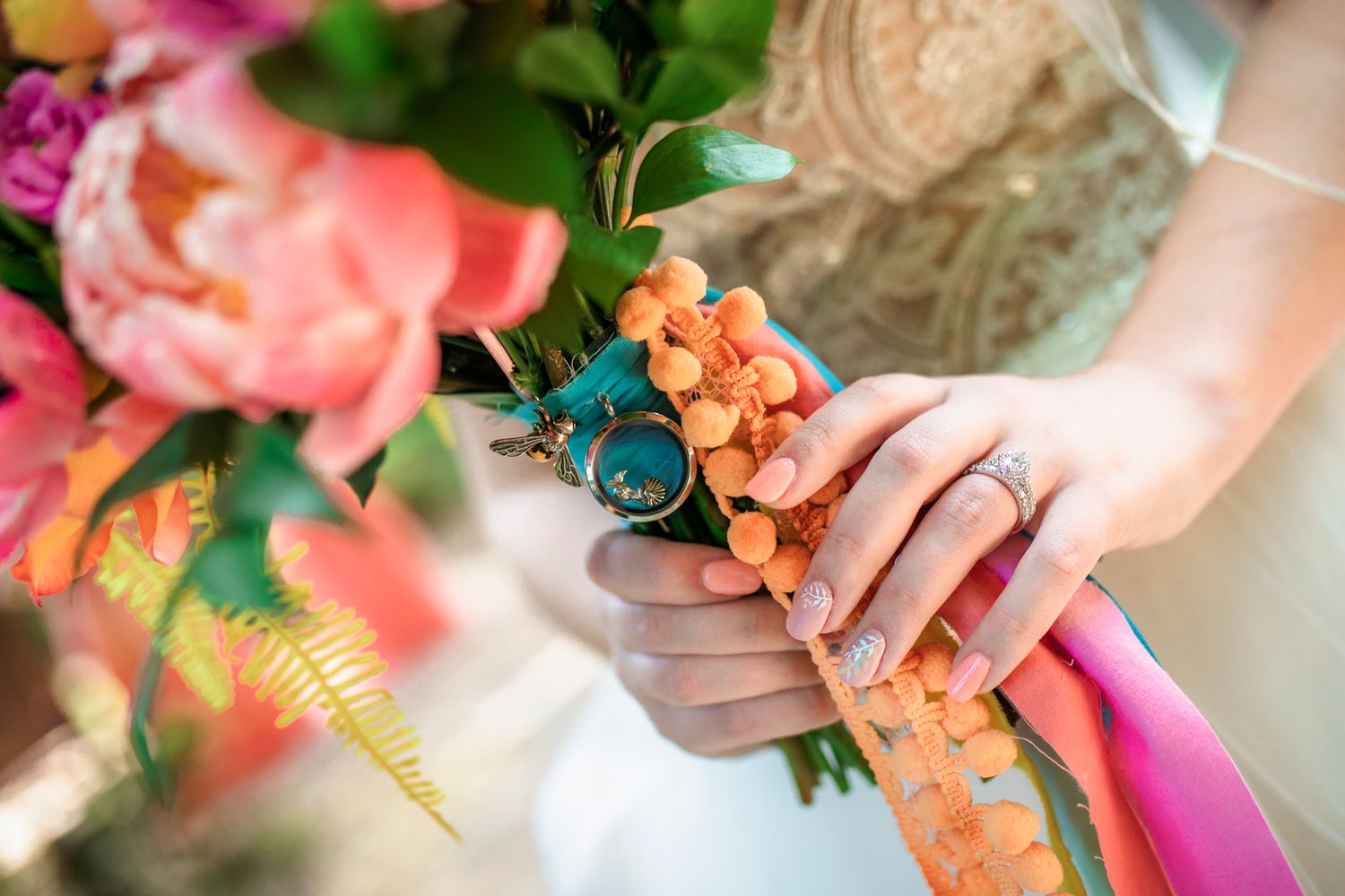 Close-up of bride holding colorful orange floral corsage with teal ribbon, wearing diamond ring, near pink peony bouquet.