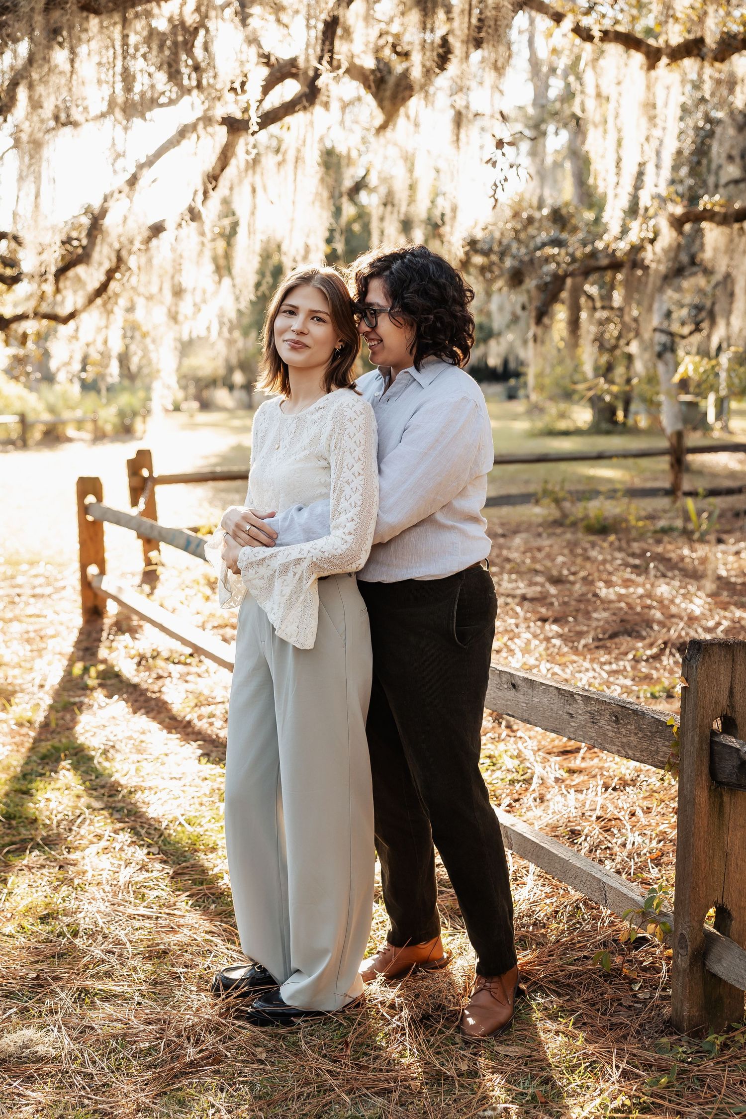 Two women standing close together under Spanish moss-draped trees near a wooden fence at golden hour.