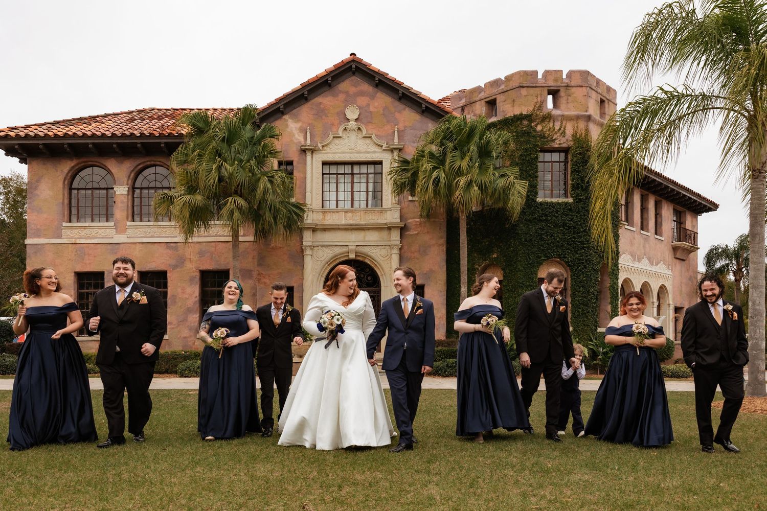 Bride and groom walk with wedding party in navy and black formal wear before an elegant historic brick building.