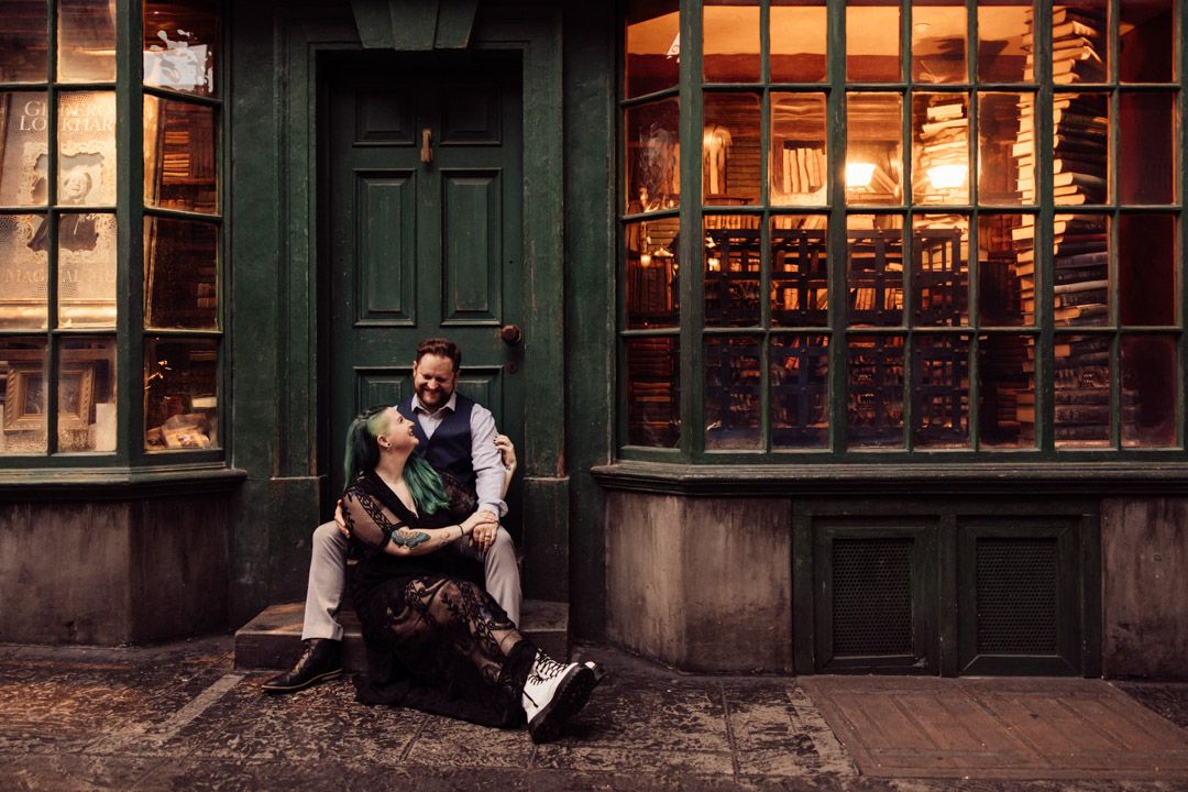 Man sitting on steps holding his partner outside a warmly lit bookshop in Diagon Alley.