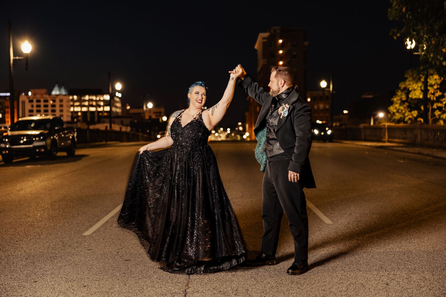 Gothic bride in black ballgown dancing with groom in street at night under city lights.