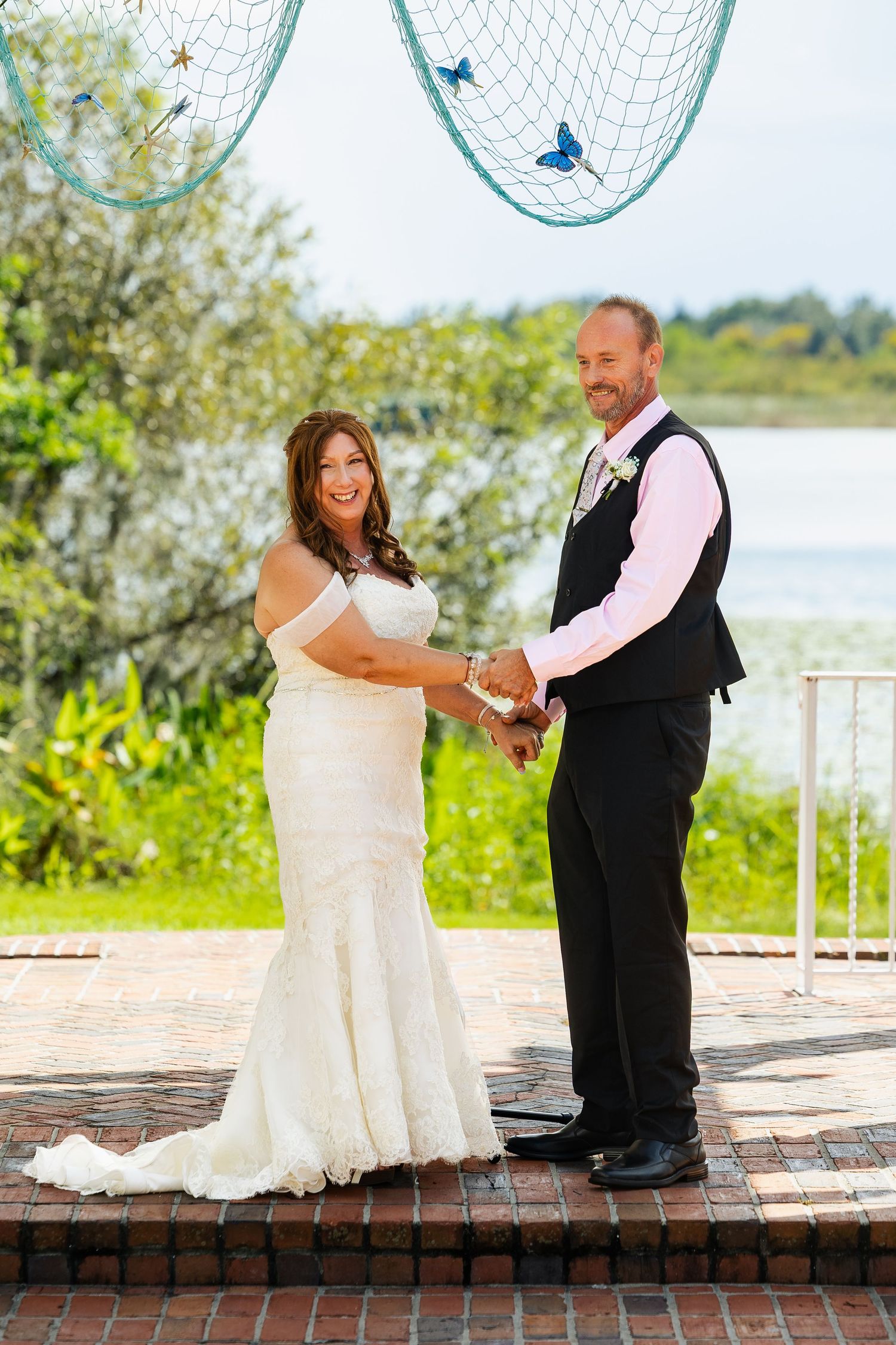 Smiling bride and groom hold hands at lakeside wedding altar decorated with butterfly net backdrop.
