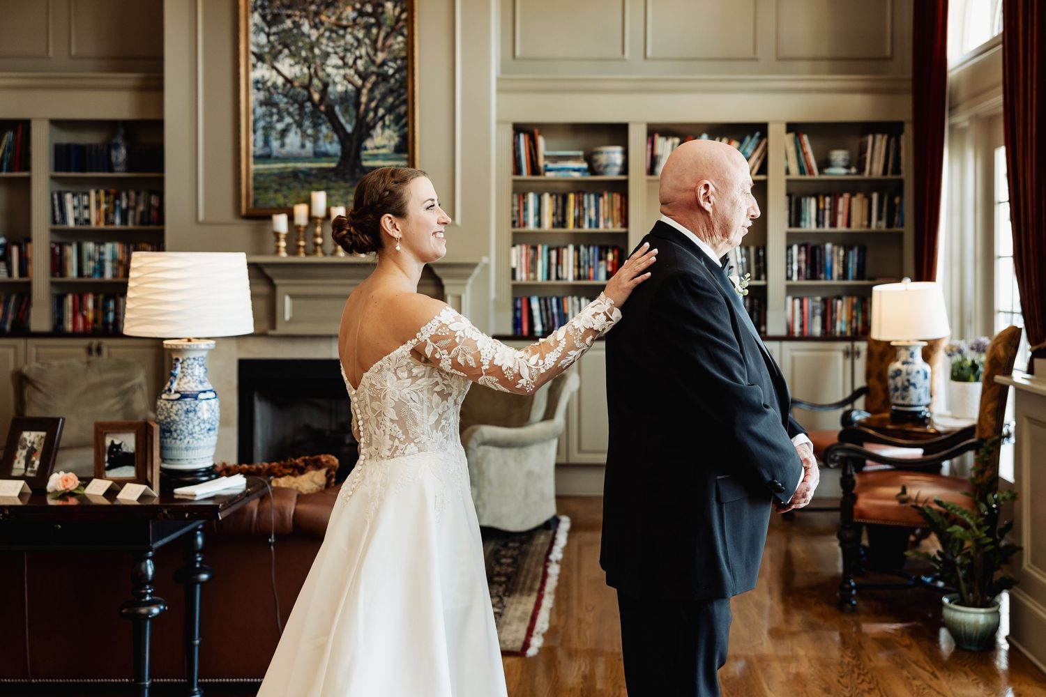 Bride in off-shoulder lace gown taps groom's shoulder during emotional wedding first look in library.