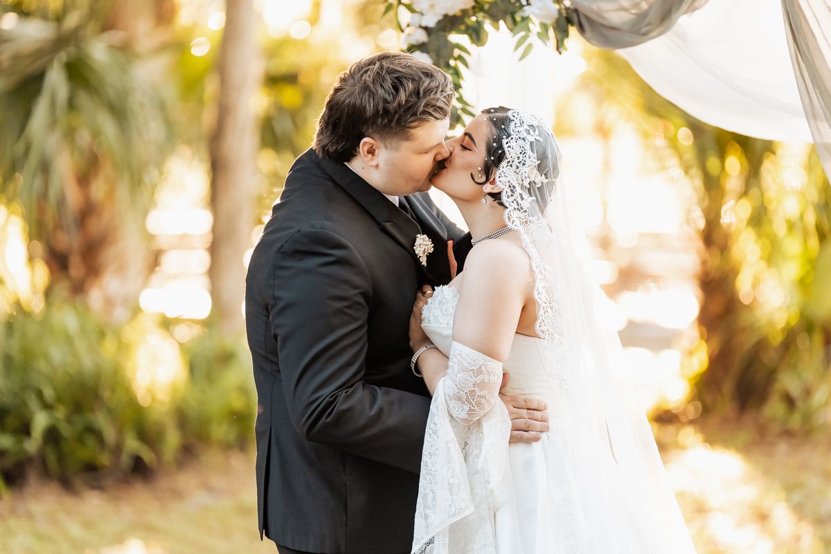 Bride and groom sharing first kiss under floral arch at outdoor wedding ceremony.