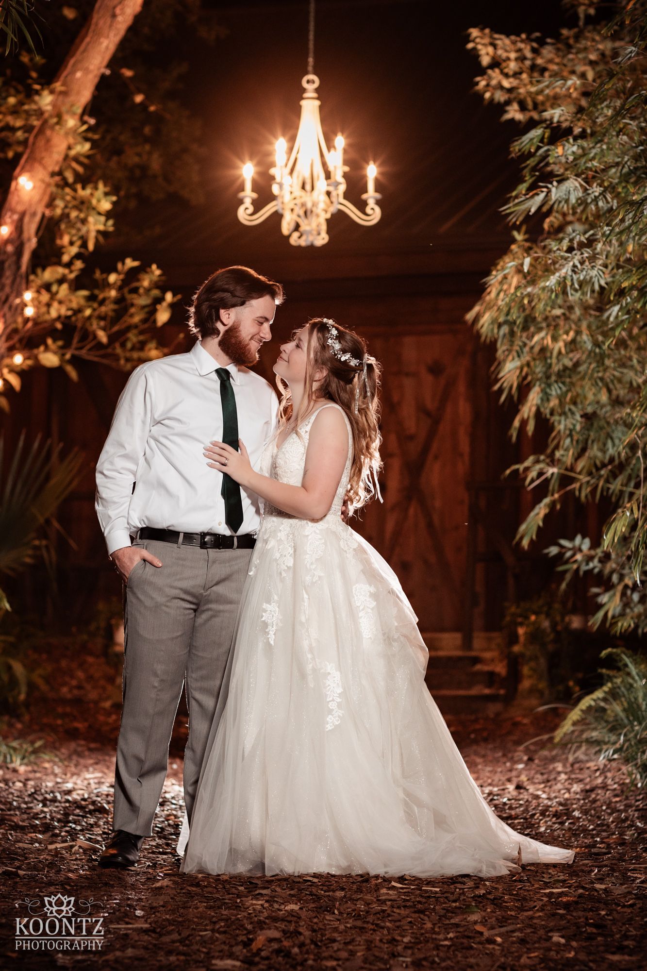 Bride and groom share romantic moment under glowing chandelier outside rustic barn wedding venue at night.