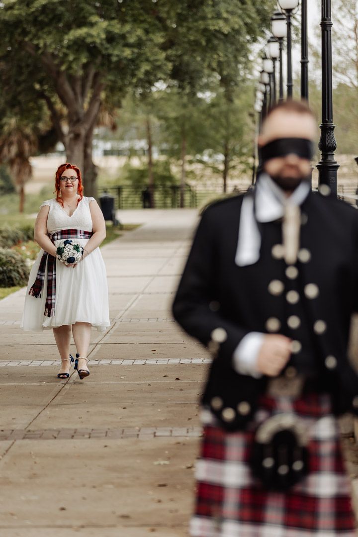 Bride in white dress with tartan sash approaches blindfolded groom in Scottish kilt on walkway.