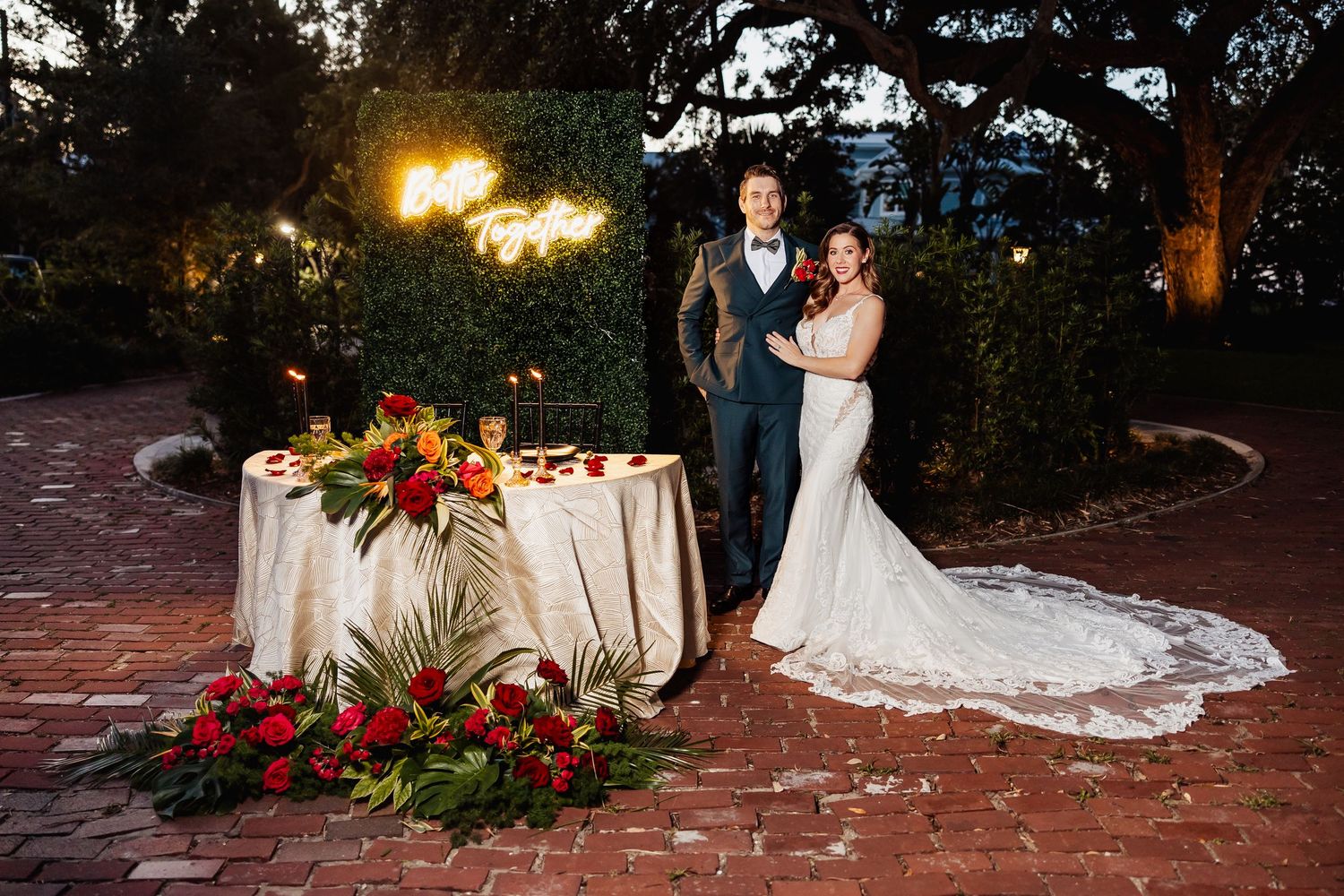 Bride and groom standing by sweetheart table with red floral arrangements.