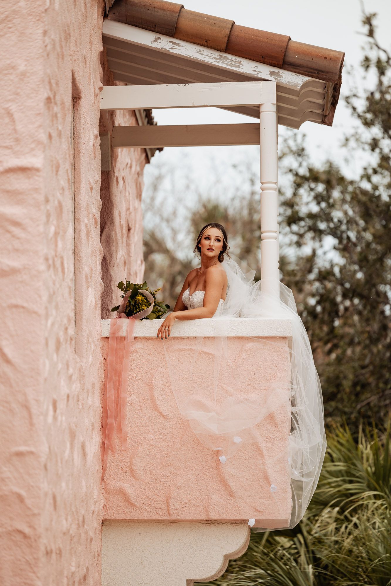 Bride in white lace gown with veil and bouquet stands on a rustic pink stucco balcony surrounded by trees.