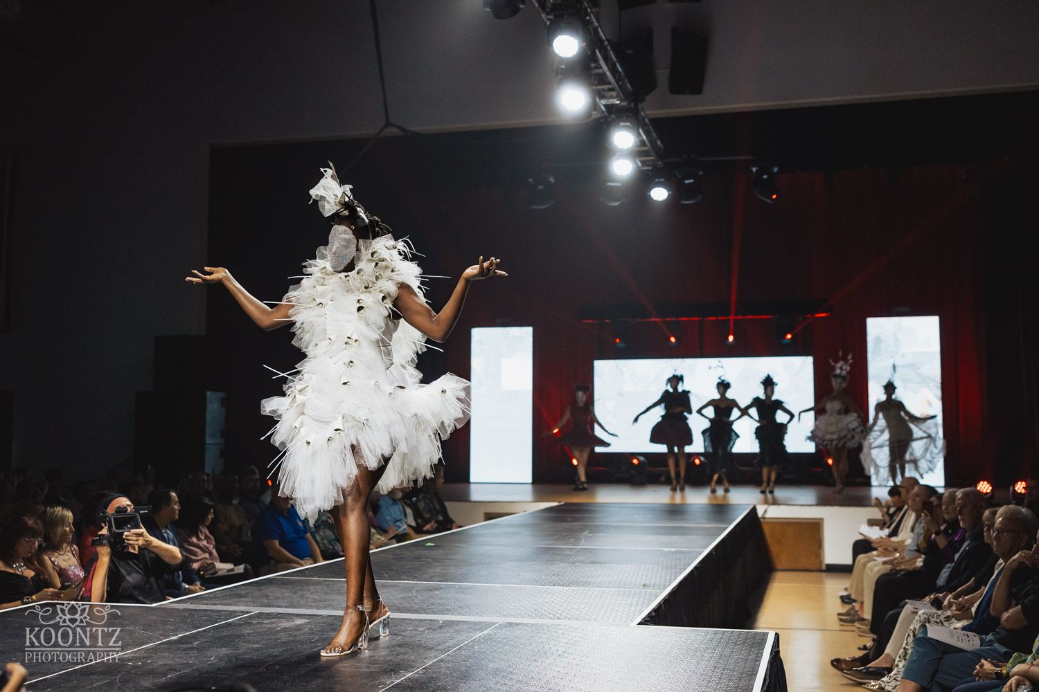 Wide view of fashion runway show with dramatic lighting and audience silhouettes visible.