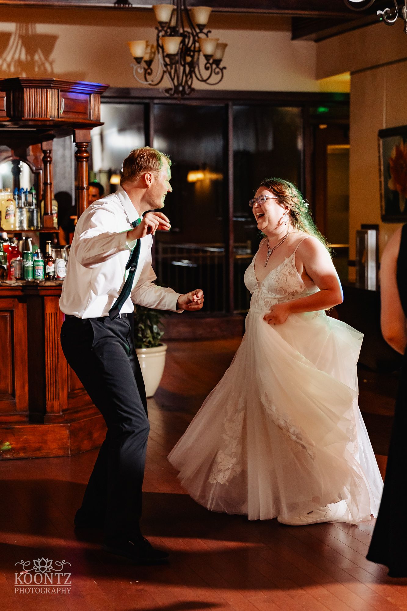 Couple dances joyfully on a wooden dance floor under an elegant chandelier at their wedding reception.