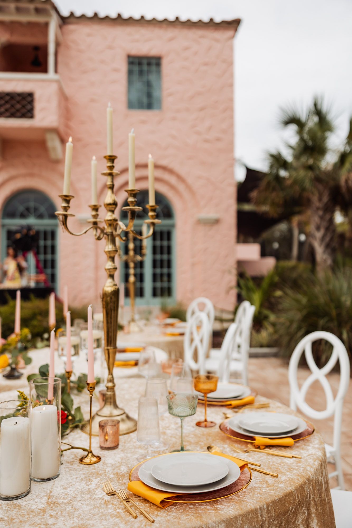 Elegant outdoor table setting with gold candelabras and white plates on sequined tablecloth against pink Mediterranean villa.