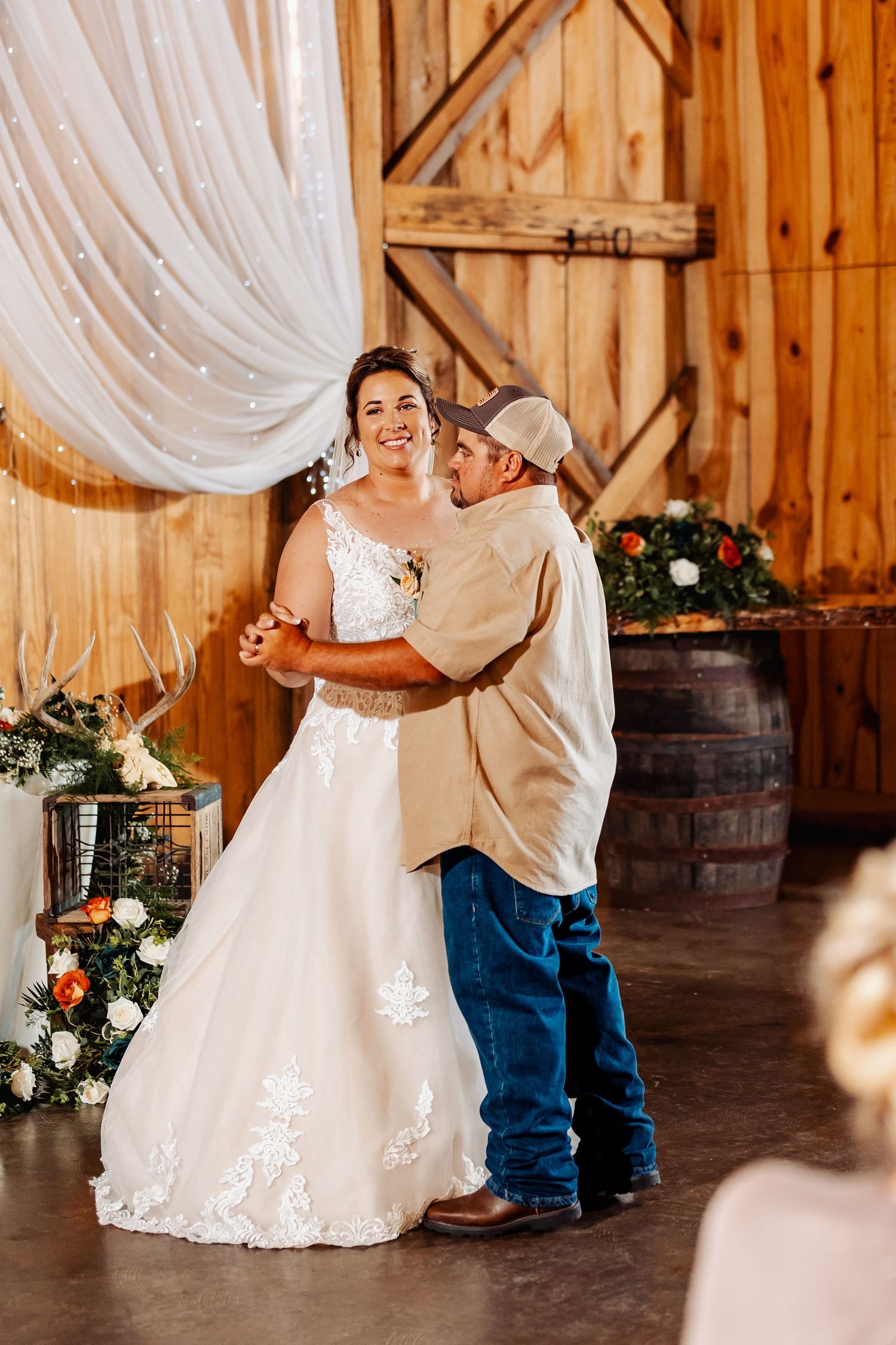 Newlyweds embrace during intimate dance moment in barn decorated with wine barrels and floral arrangements.