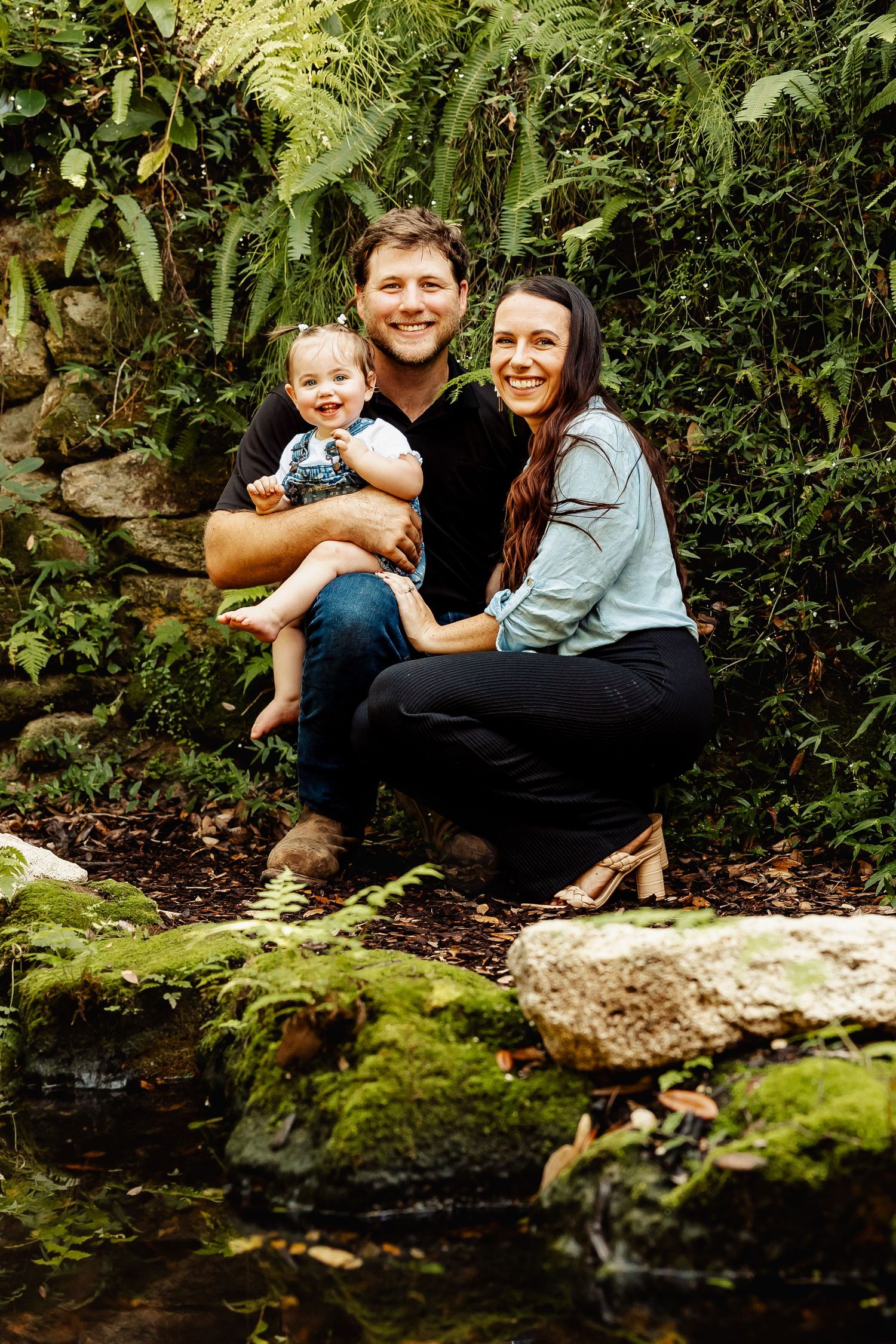 Happy family with baby posing among lush green ferns and mossy rocks in a natural outdoor setting.