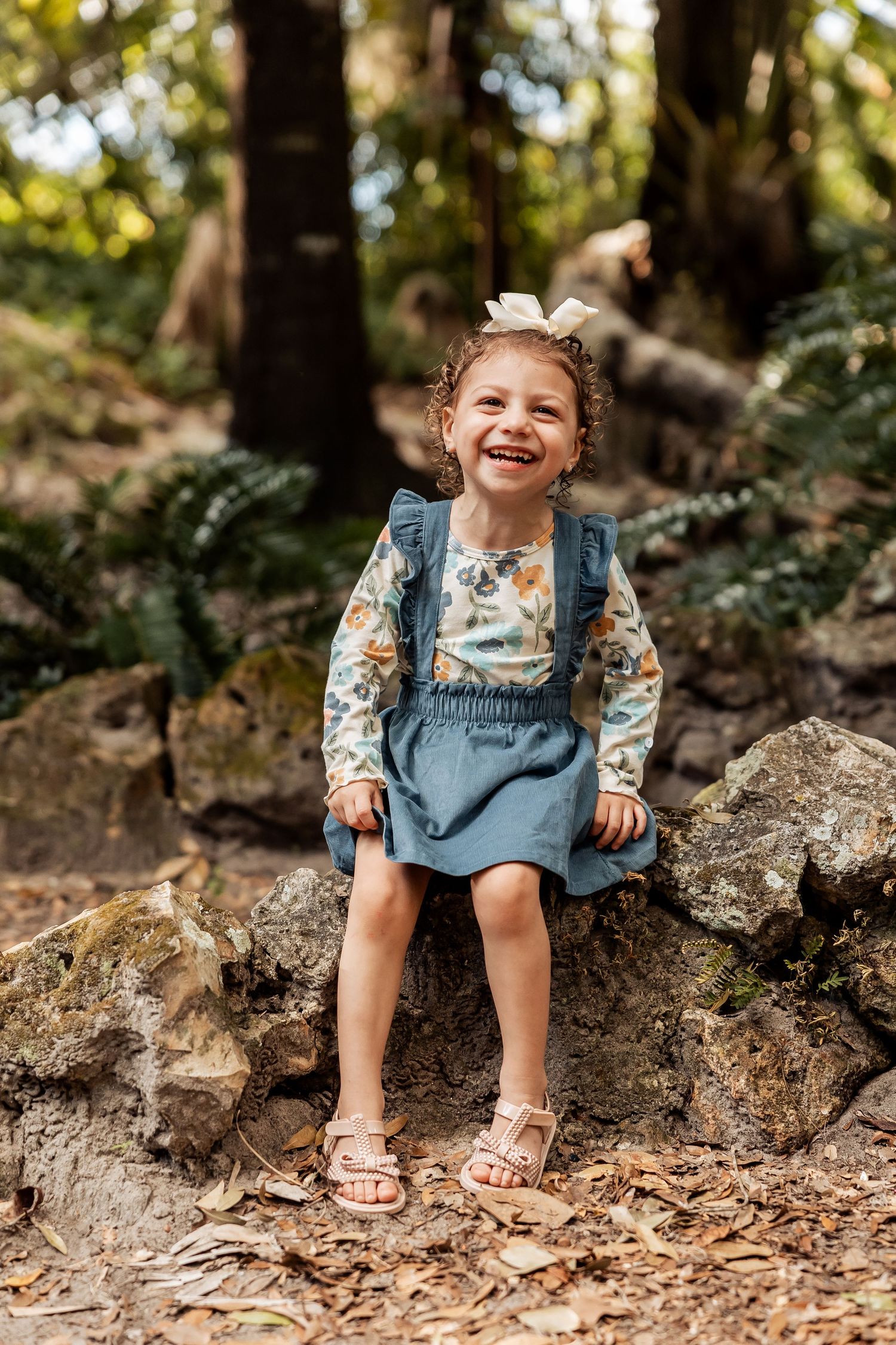 A joyful curly-haired toddler girl in a floral outfit laughs while sitting on a mossy rock outdoors.