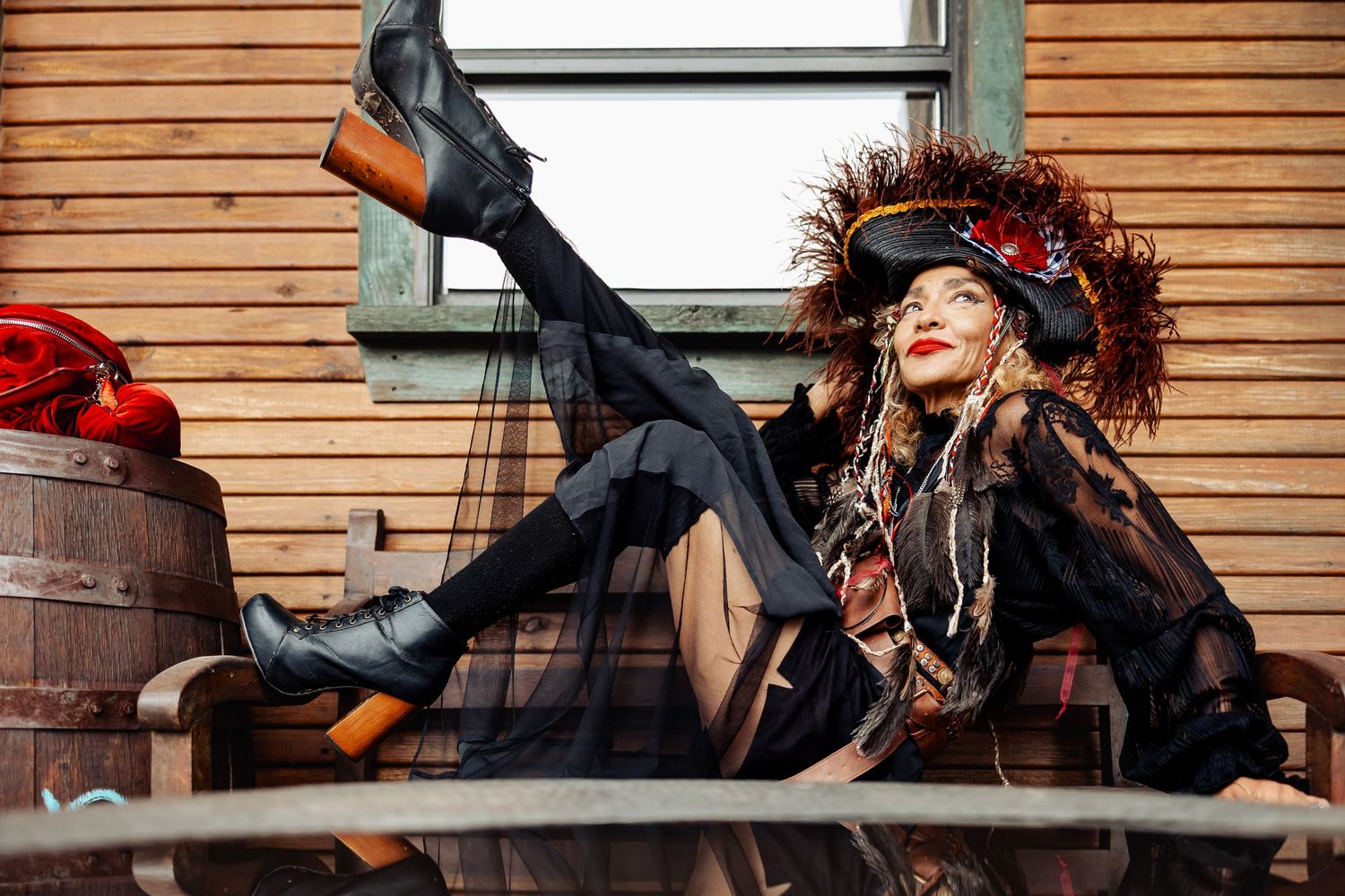 Woman in pirate costume with feathered hat poses dramatically on wooden bench near barrel.