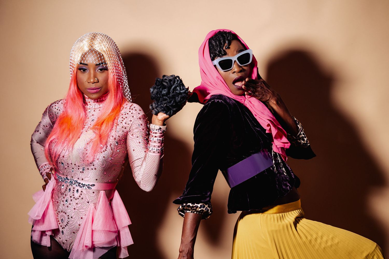 Two women in vibrant pink wigs and bold fashion outfits pose together against a neutral beige background.