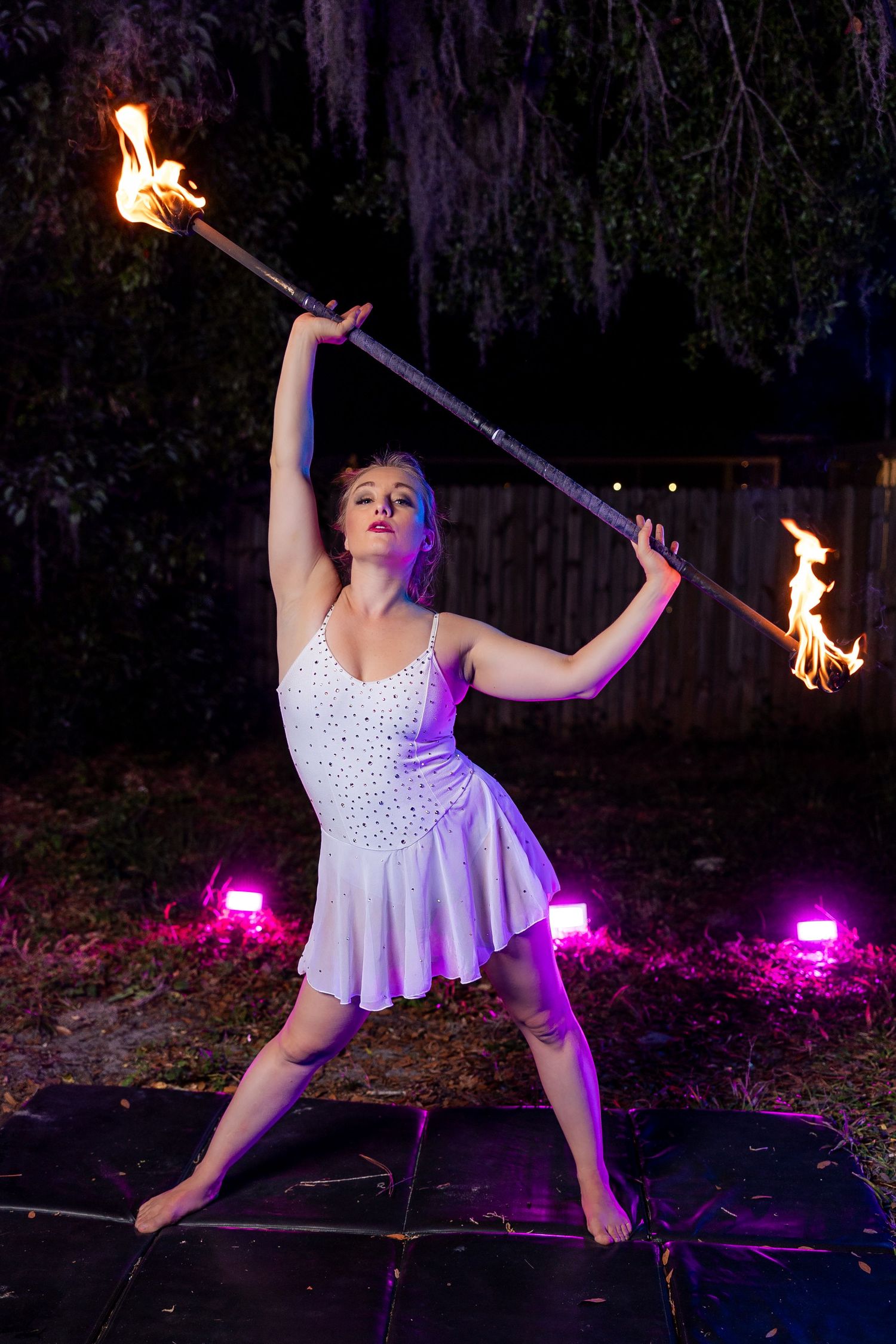 Barefoot female fire performer in white dress posing with a lit double-ended fire staff at night.