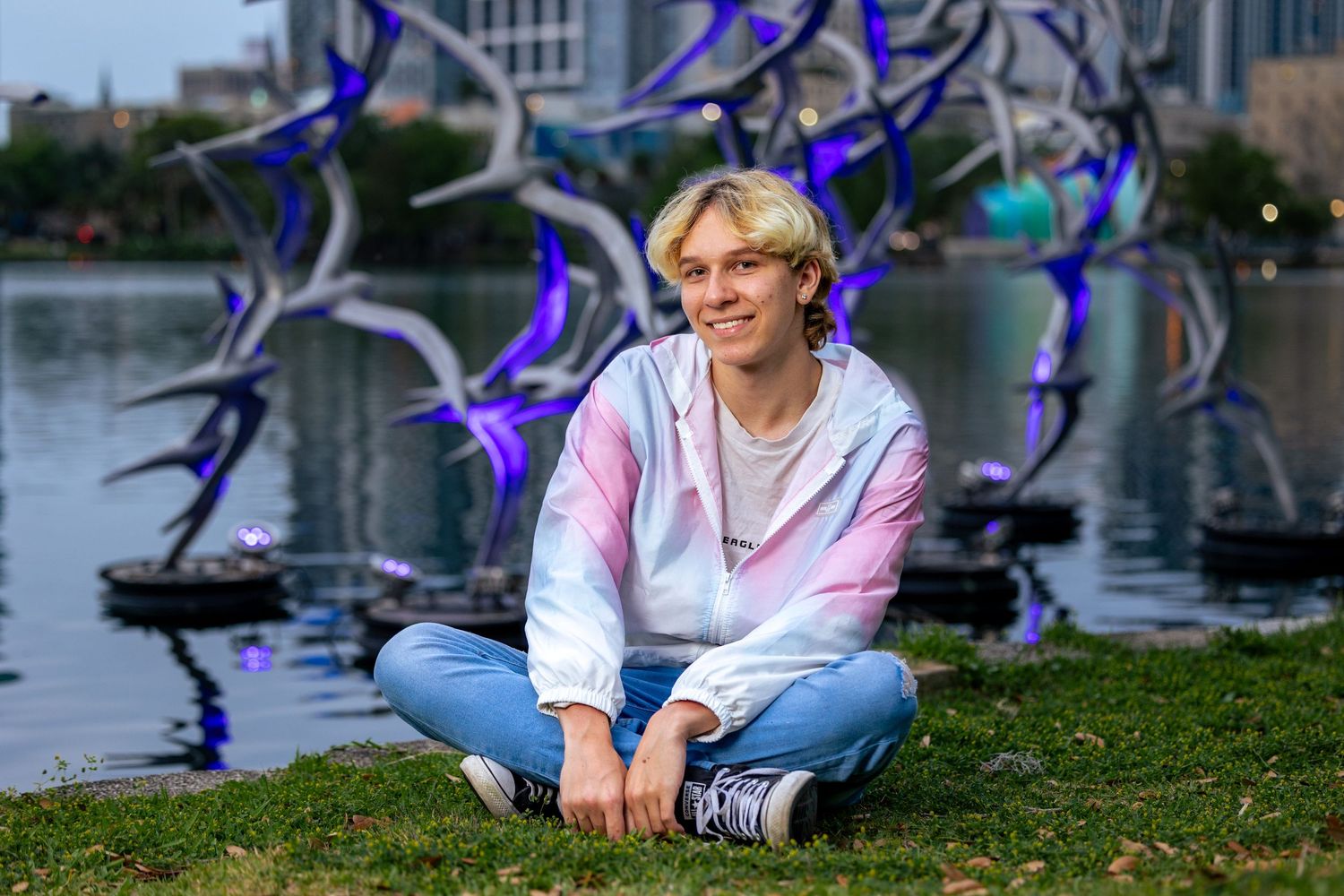 Teen boy in pink hoodie sits cross-legged on grass by lake with blue metal bird sculptures.