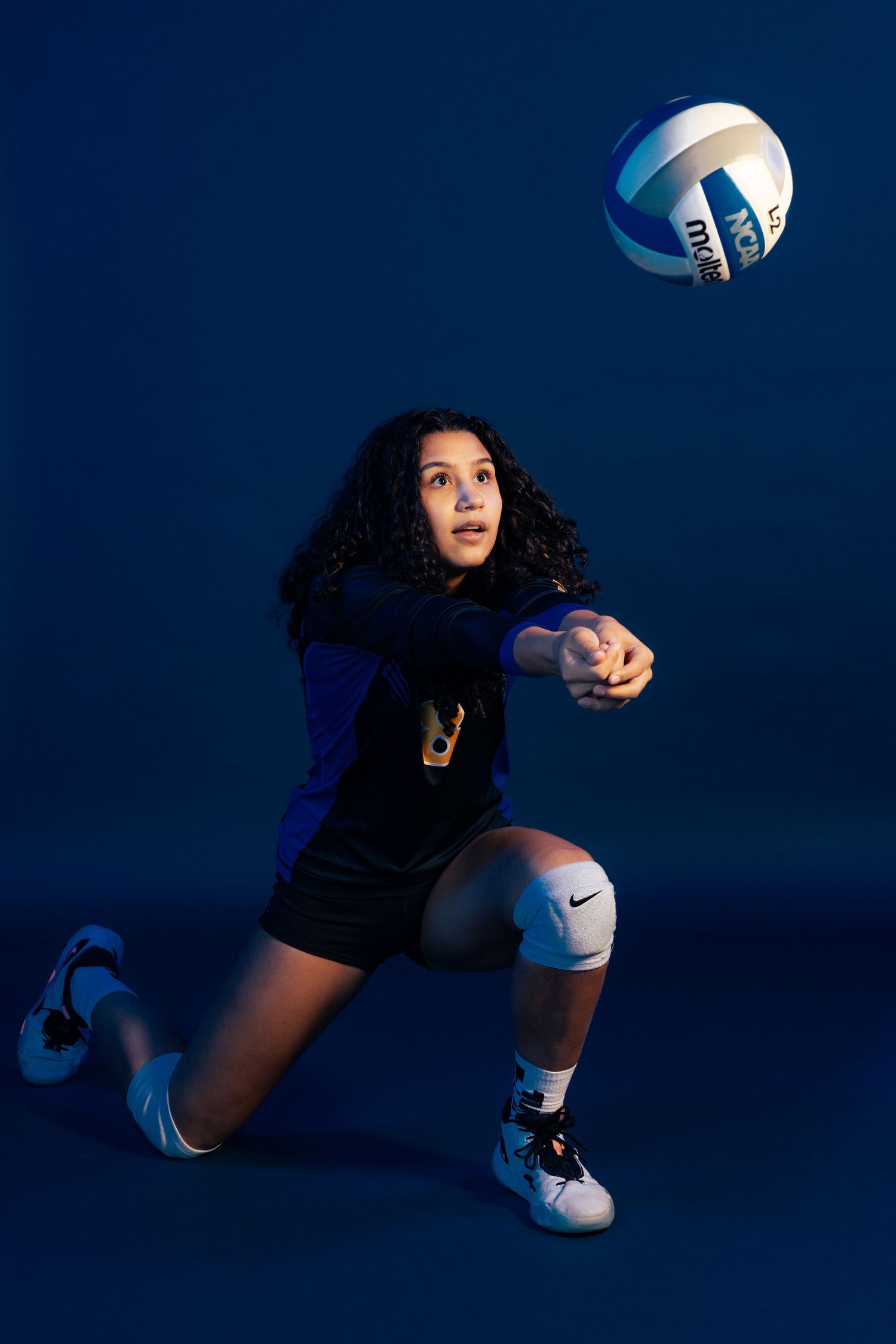 Female volleyball player in blue uniform performing a dig, bumping the ball in a dramatic studio shot.