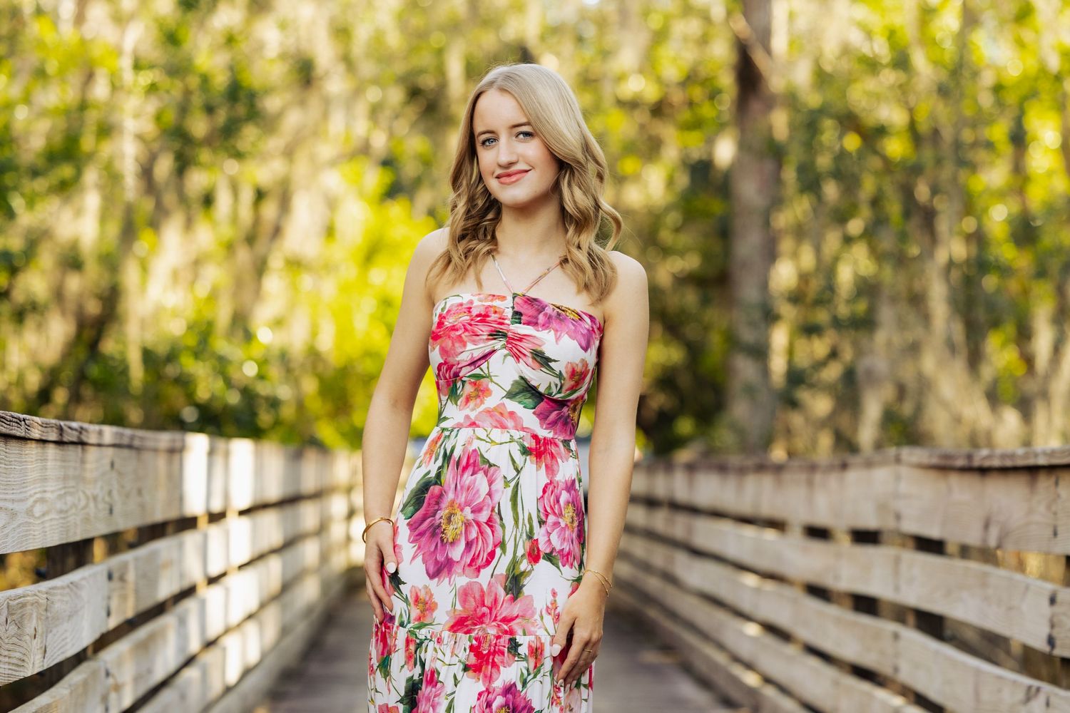 Young woman in floral strapless dress smiling on wooden bridge surrounded by lush green trees.